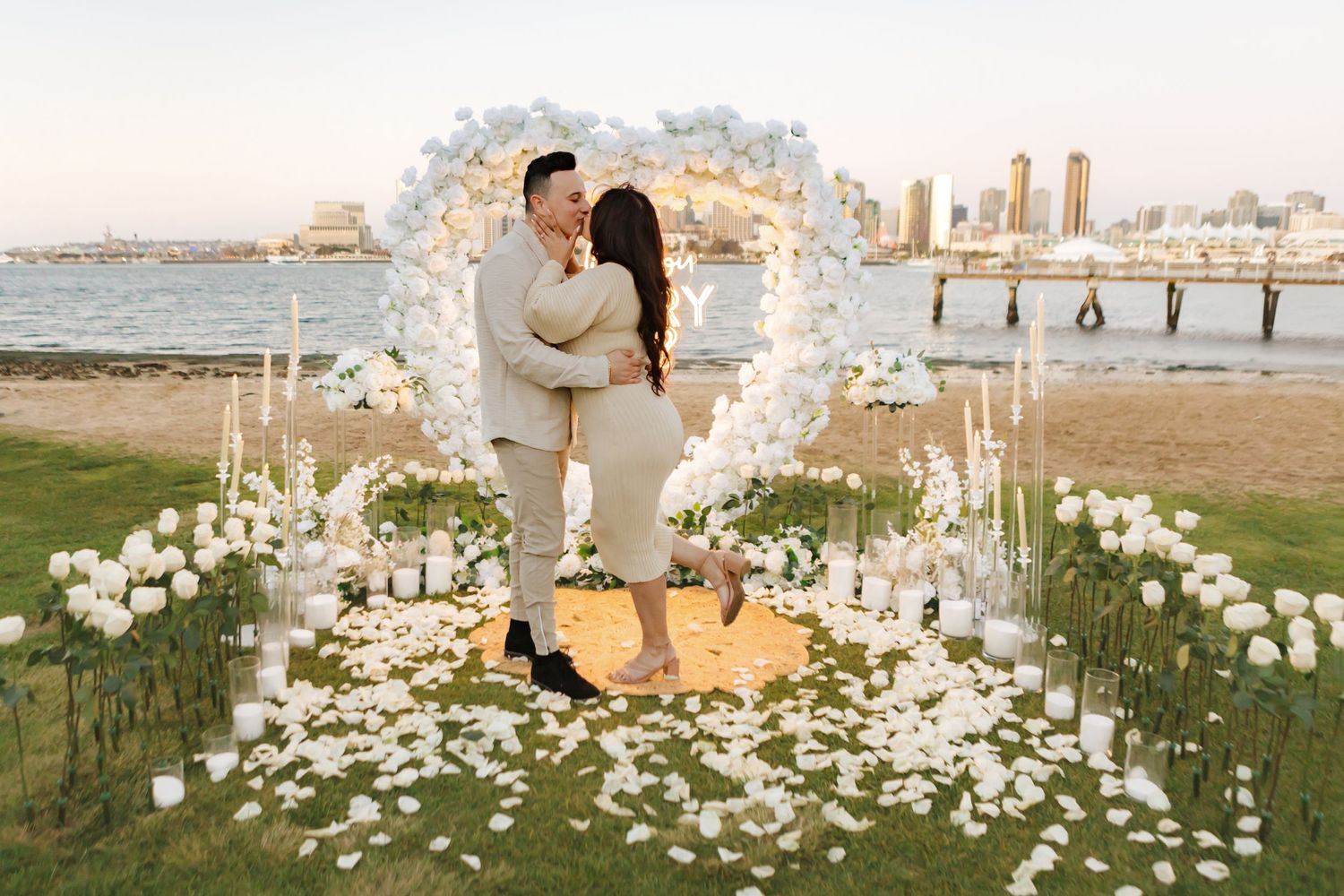 Couple embraces under white floral arch with cityscape backdrop during romantic proposal.