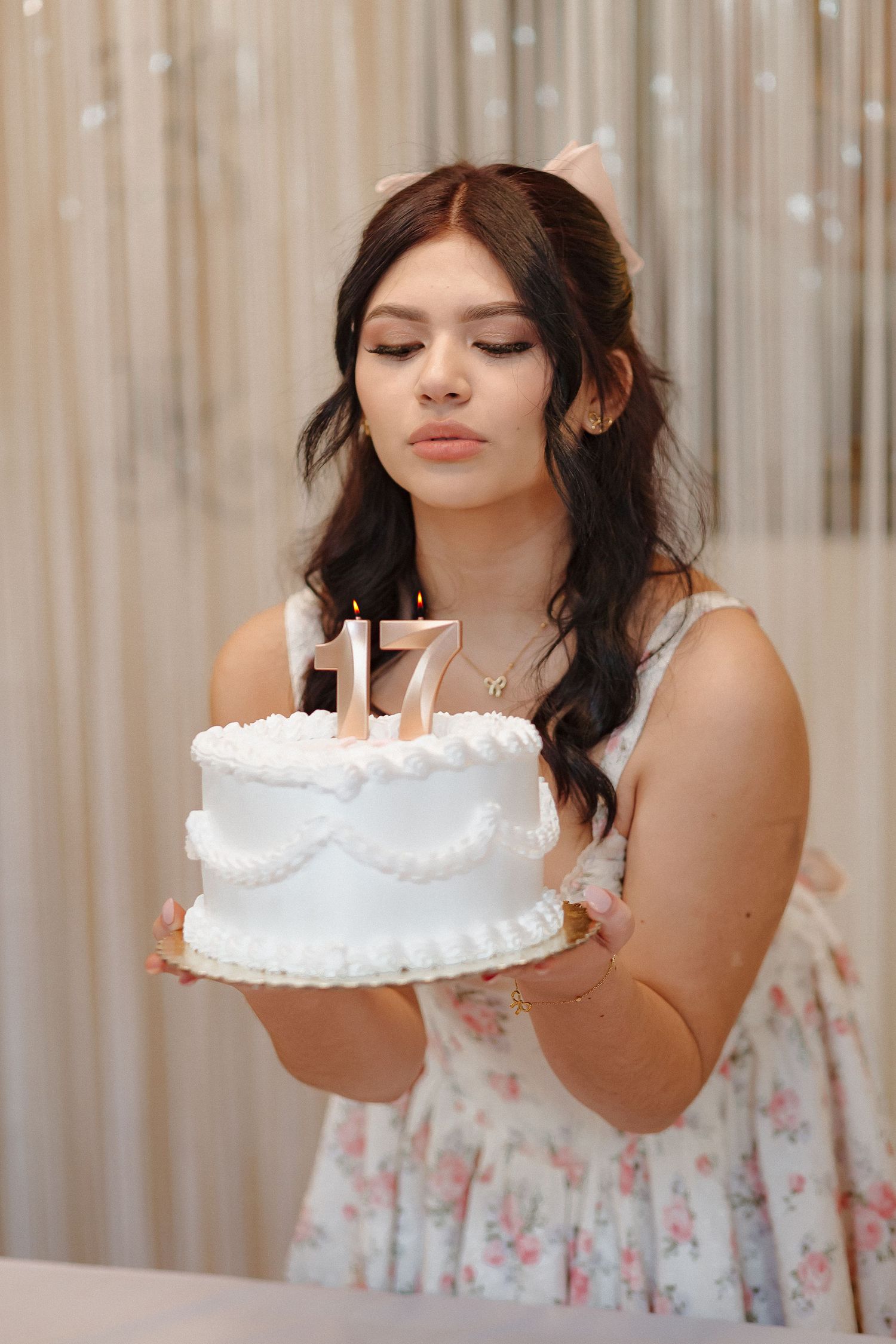 White birthday cake with lit candles displayed against shimmering curtain backdrop.