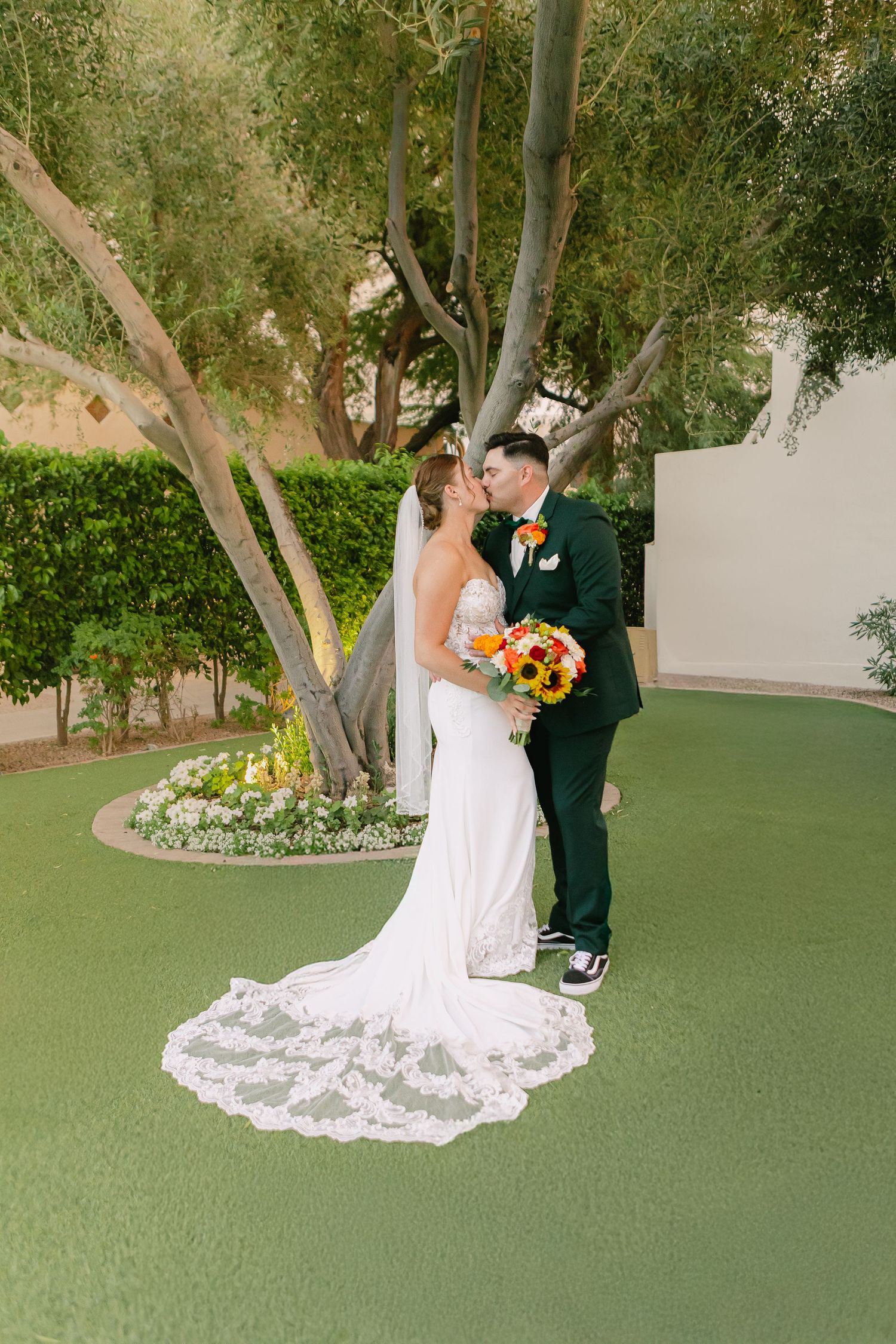 A wedding couple poses beneath trees on a green lawn with sunflowers in their formal attire.