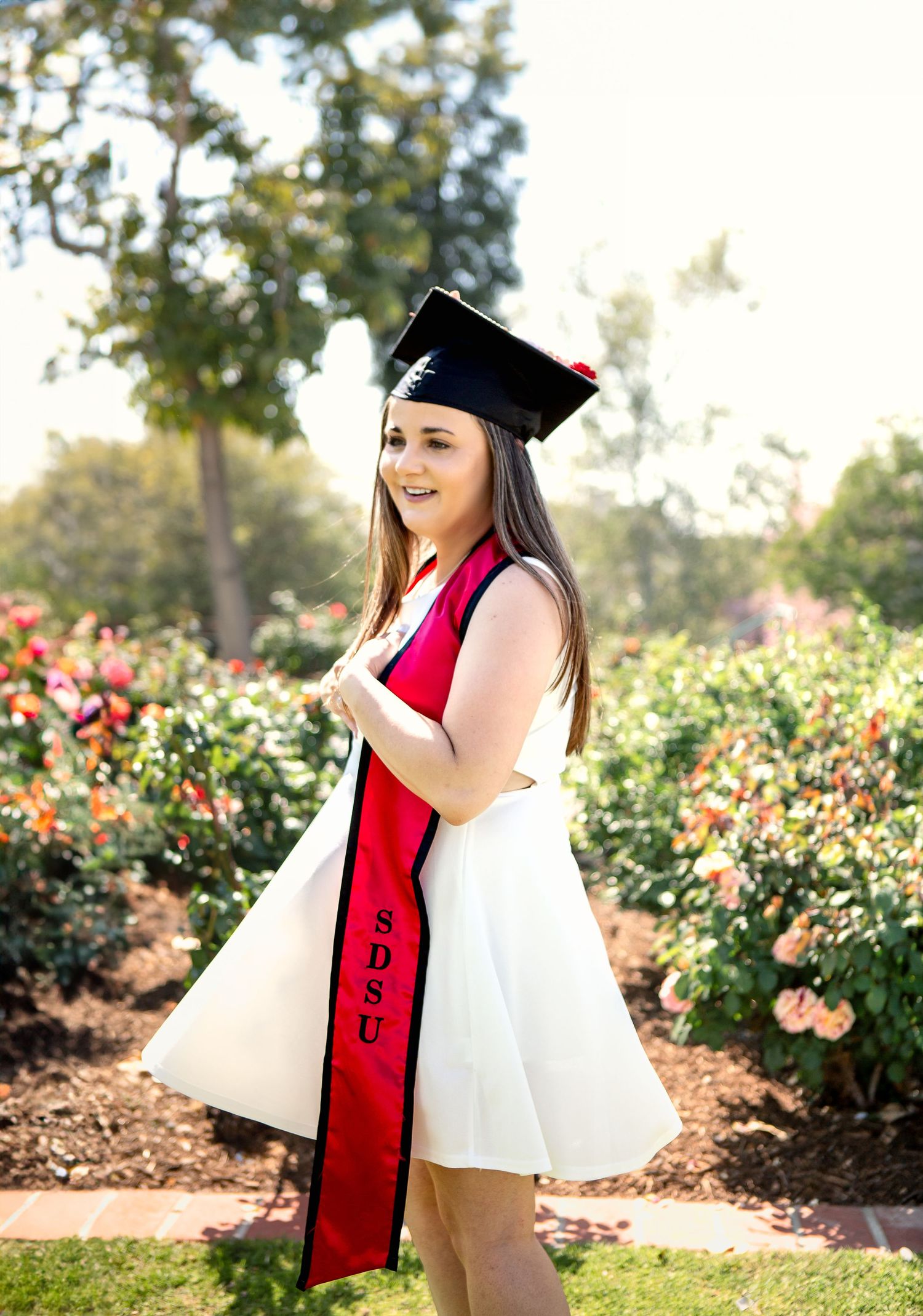 Graduate in white dress and red stole poses in garden with blooming flowers.