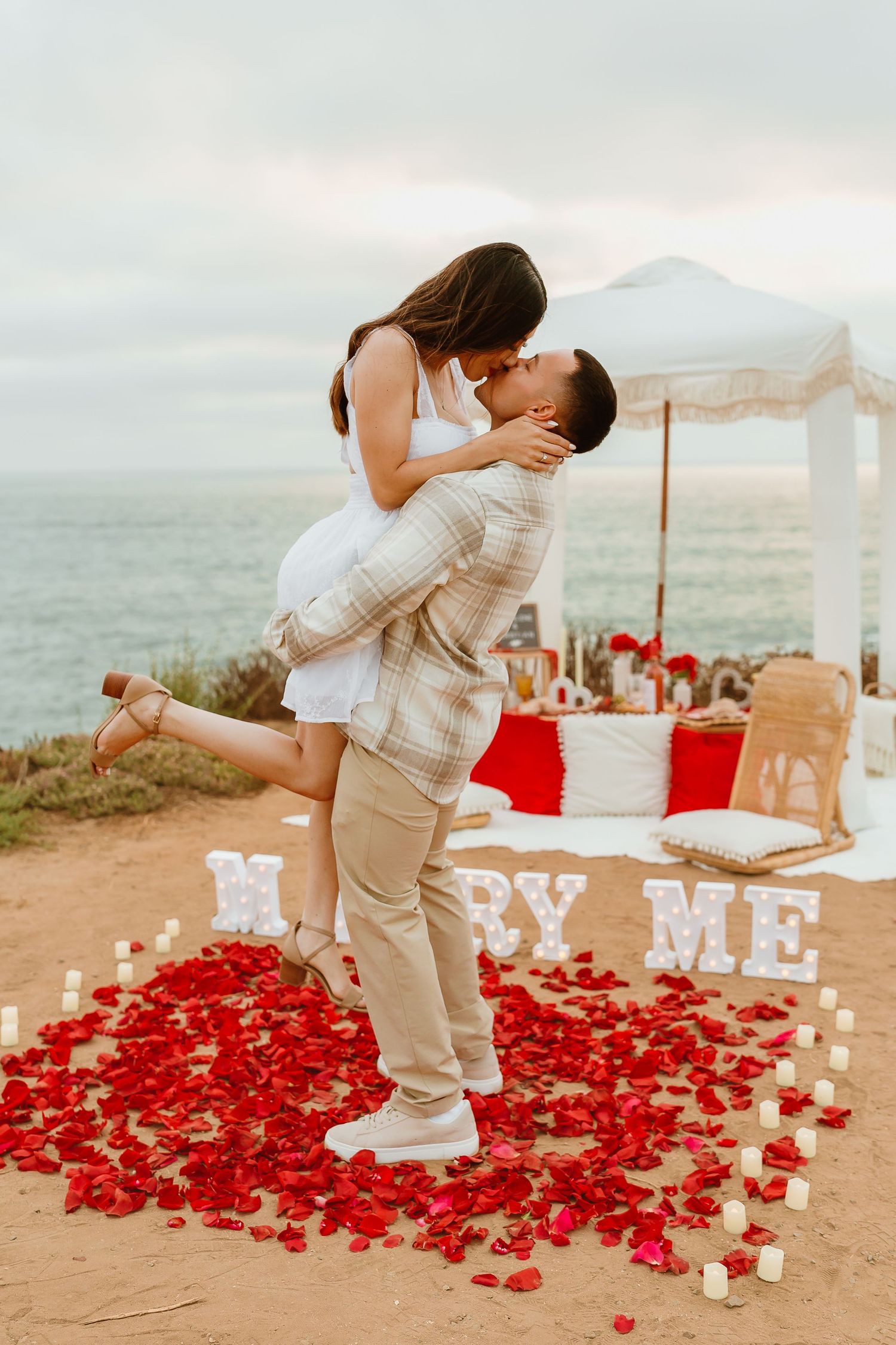 Romantic proposal setup with candles and rose petals spelling 'Marry Me' at sunset.