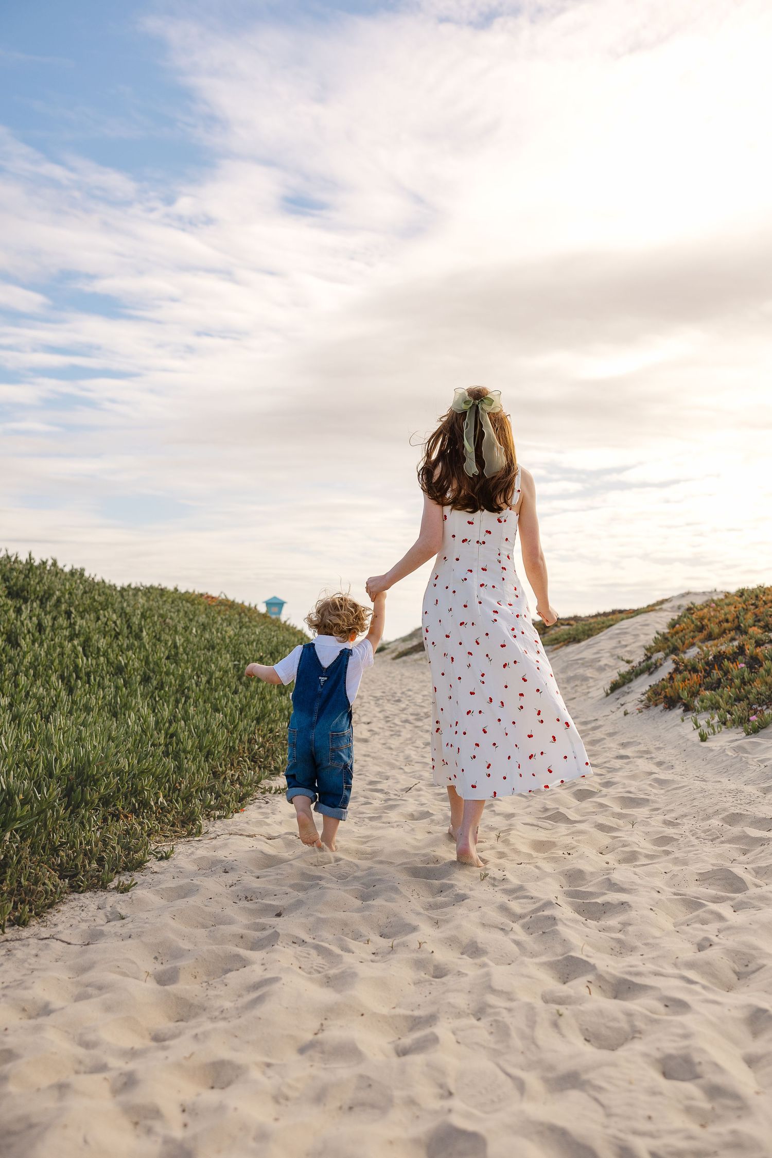 Two figures walk hand in hand up a sandy beach path bordered by green grass on a cloudy summer day.