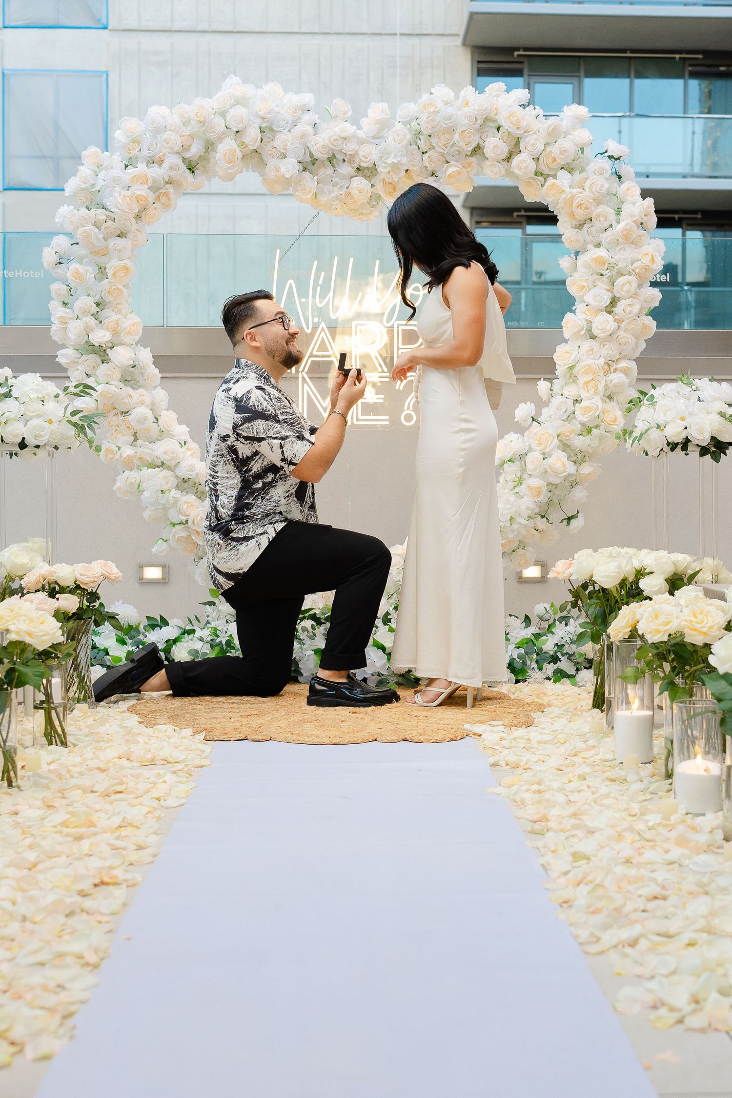 A proposal scene under a white floral arch with candles and rose petals.