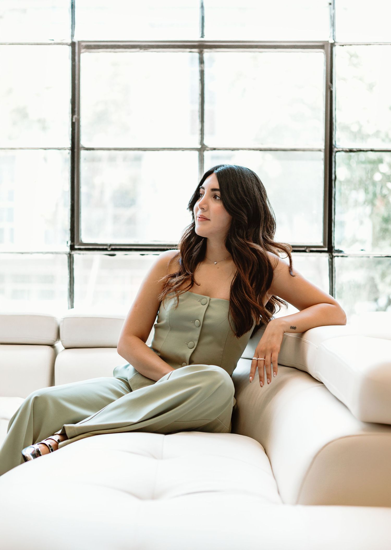 Casual portrait of person relaxing on white couch near bright window wearing green button-up dress.