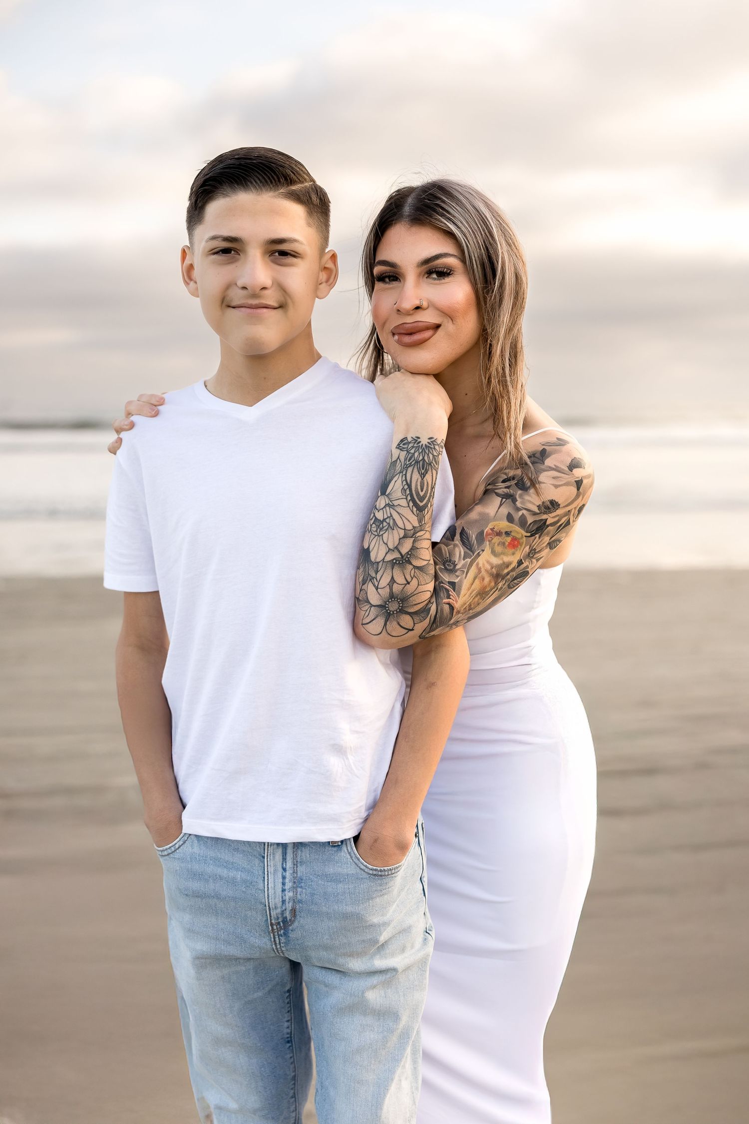 A casual portrait of a pair in white outfits standing on a beach at sunset, one with decorative arm tattoos.