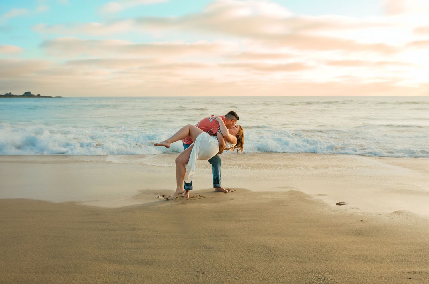 Playful beach engagement photo with couple posing against sunset ocean backdrop.