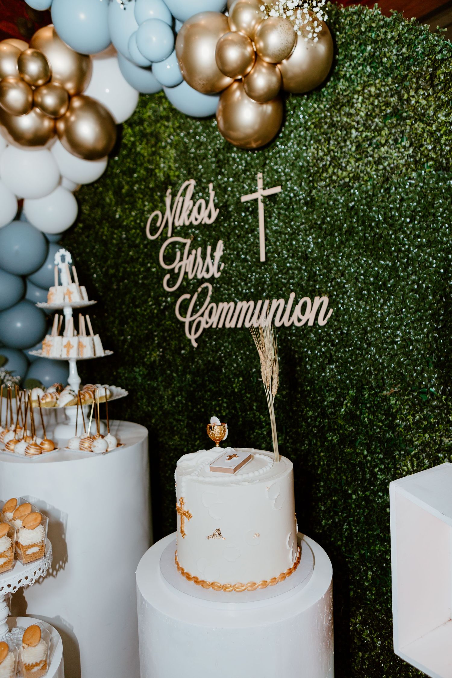 First Communion celebration setup with white cake, blue and gold balloons against green hedge wall.