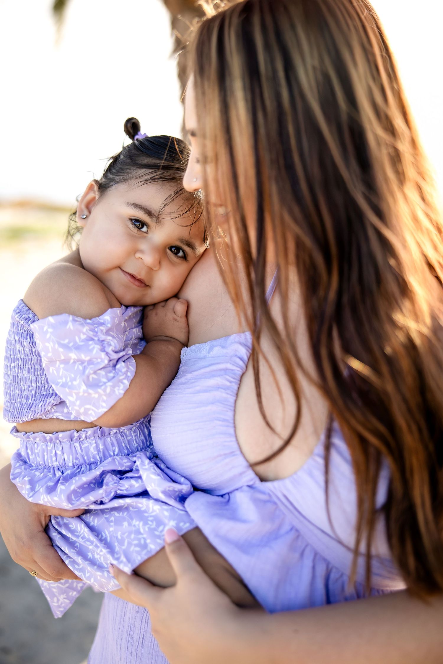Adorable toddler in purple lace dress cuddles with parent during outdoor sunset photoshoot.