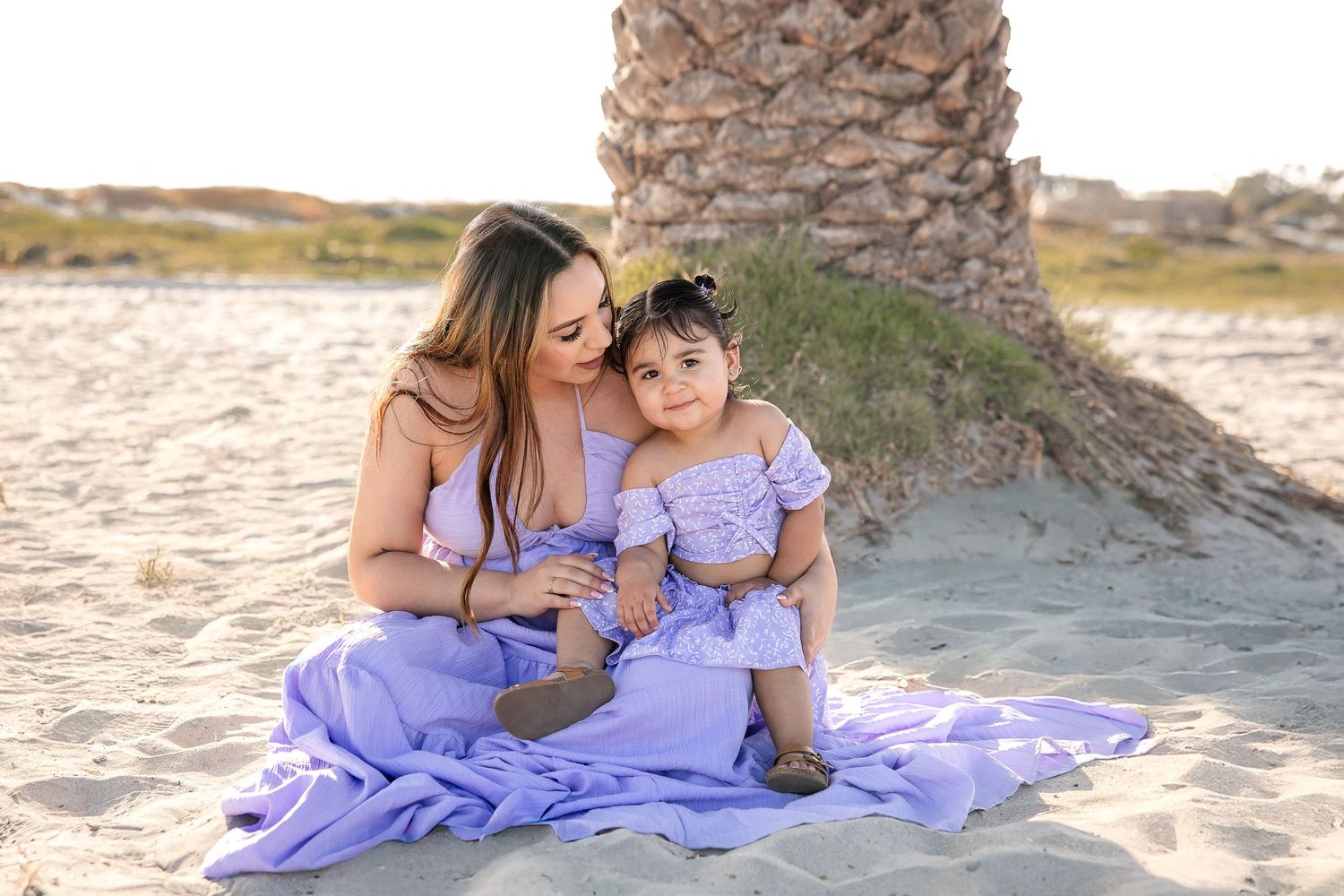 Two people in matching lilac dresses sit together on a blanket at the beach near a palm tree at sunset.