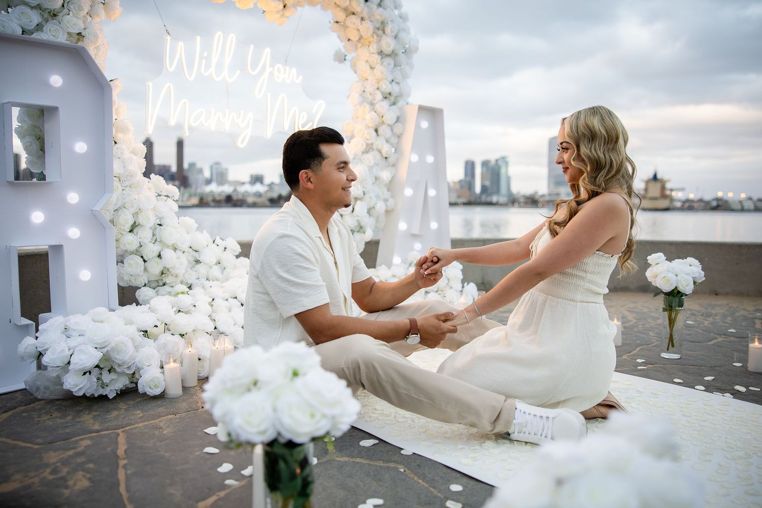 A romantic rooftop proposal scene decorated with white flowers and candles against a cityscape backdrop during sunset.