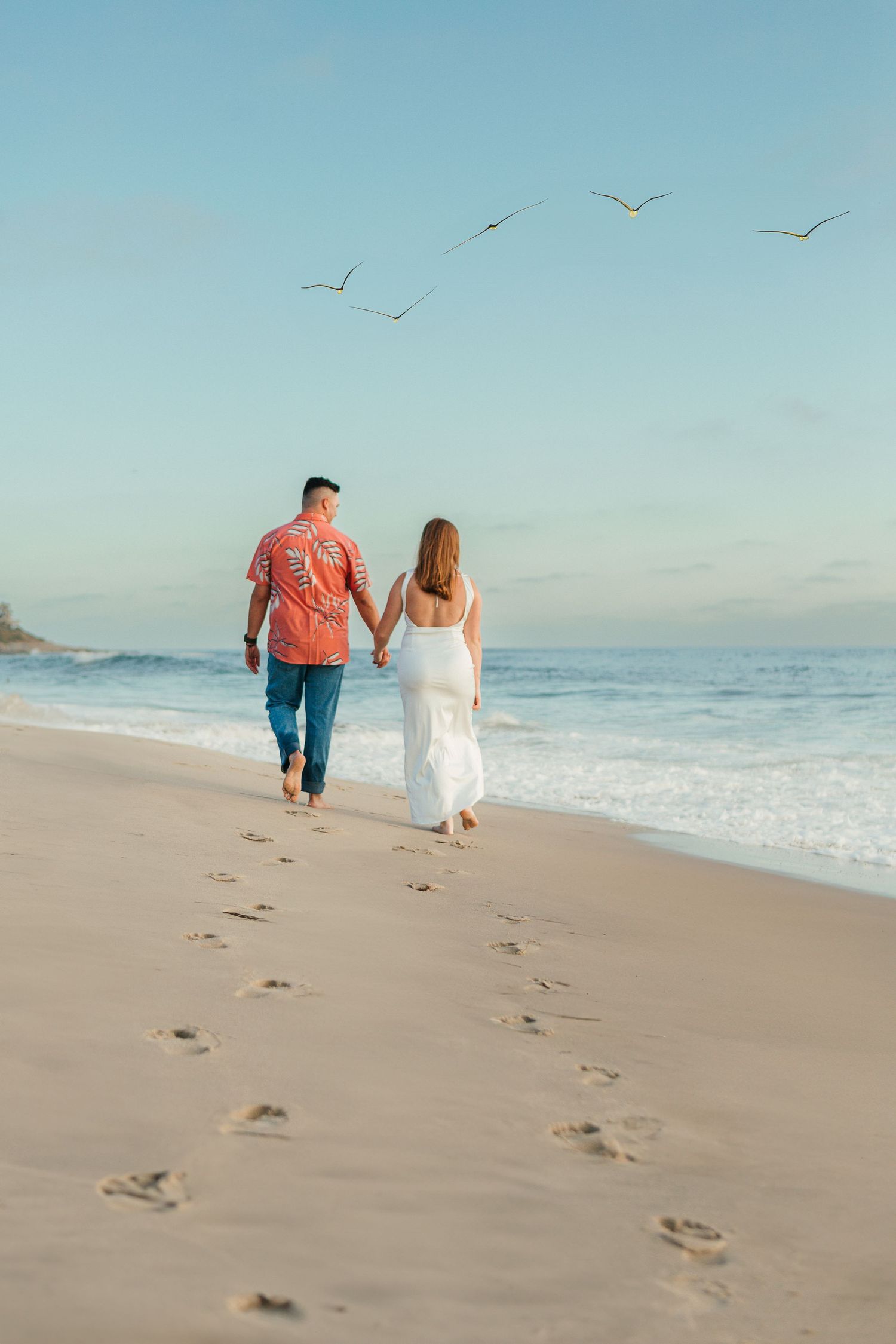 Couple walking on beach leaving footprints in sand with seagulls flying overhead.