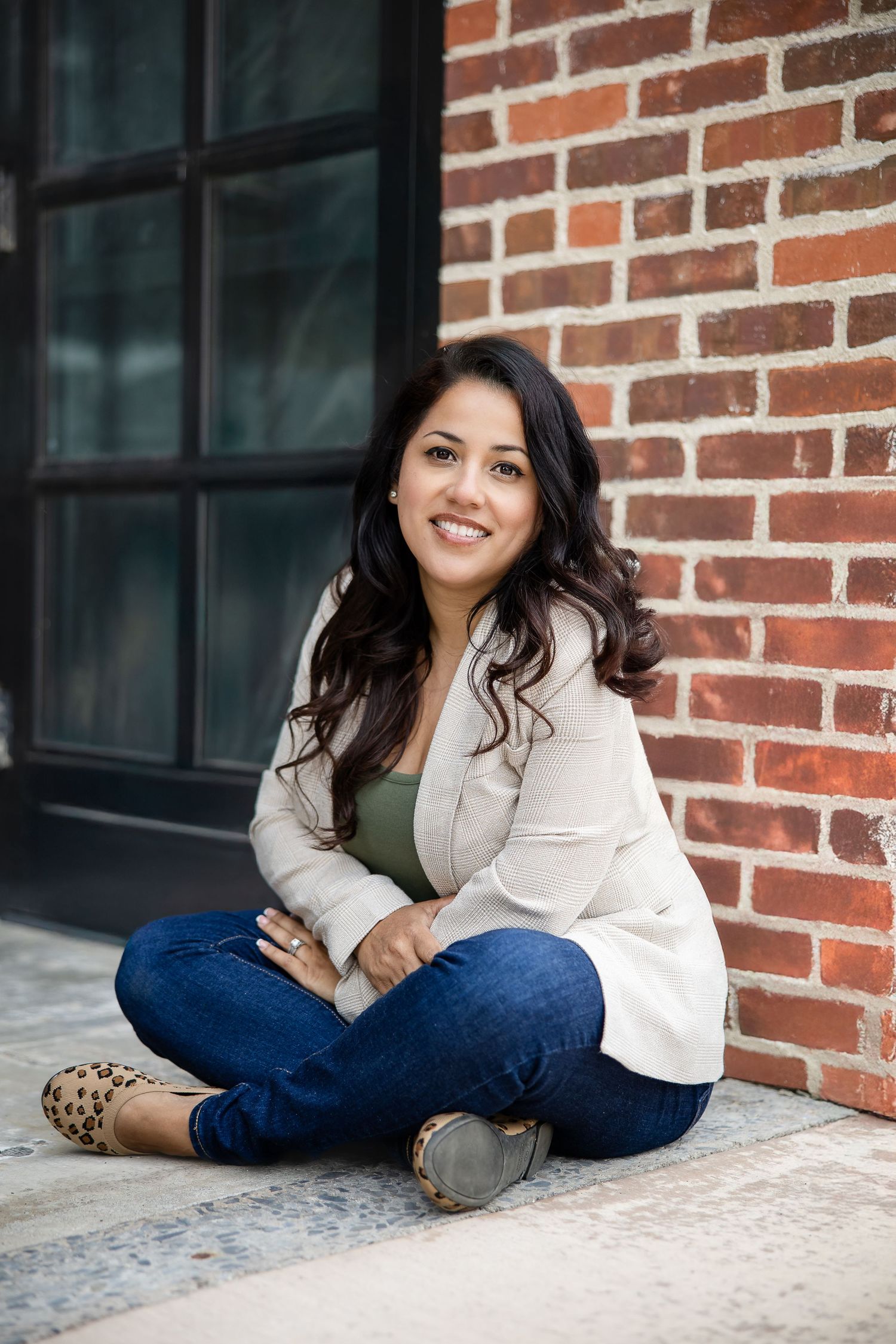 Professional lifestyle portrait of someone in a cream blazer and jeans sitting casually against a brick wall exterior.
