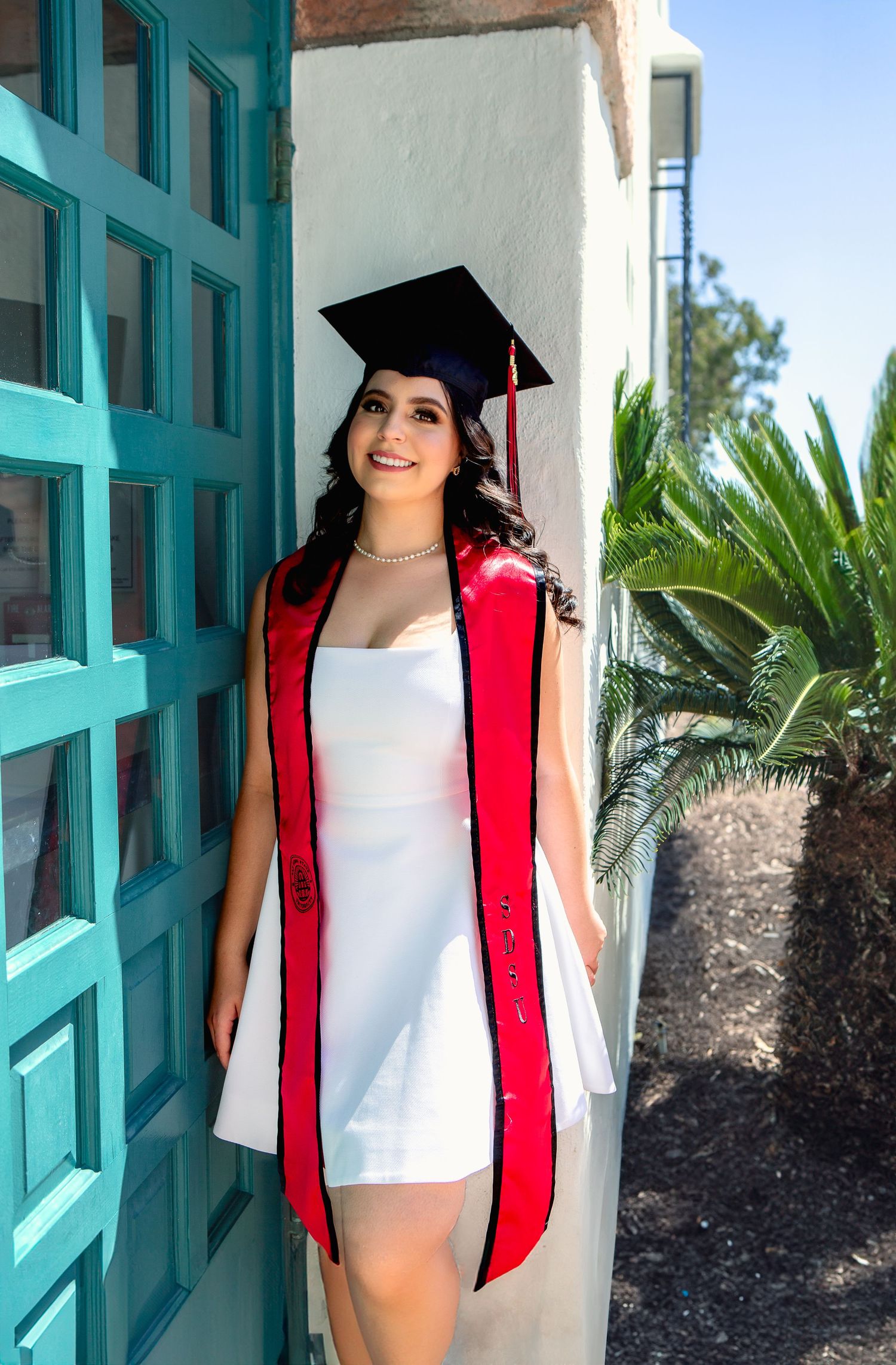 Graduate in white dress and red stole poses by turquoise door during graduation photoshoot.