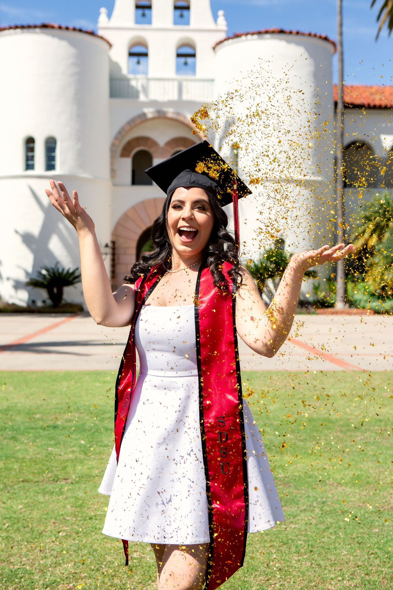 Graduate celebrating with glitter toss in front of white Spanish-style building.
