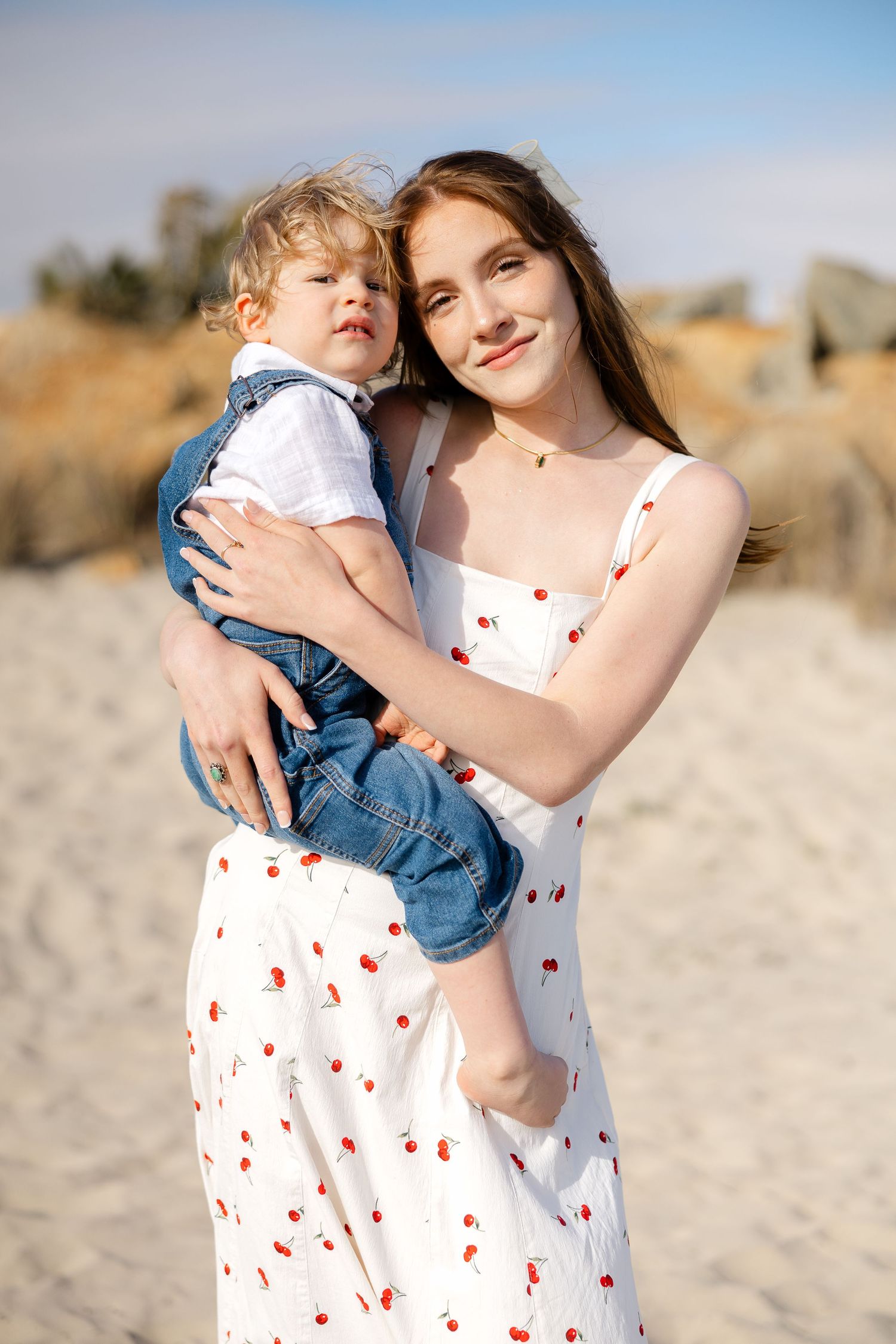 A sweet outdoor portrait shows a parent and child embracing at the beach, wearing a polka dot dress and casual clothes.