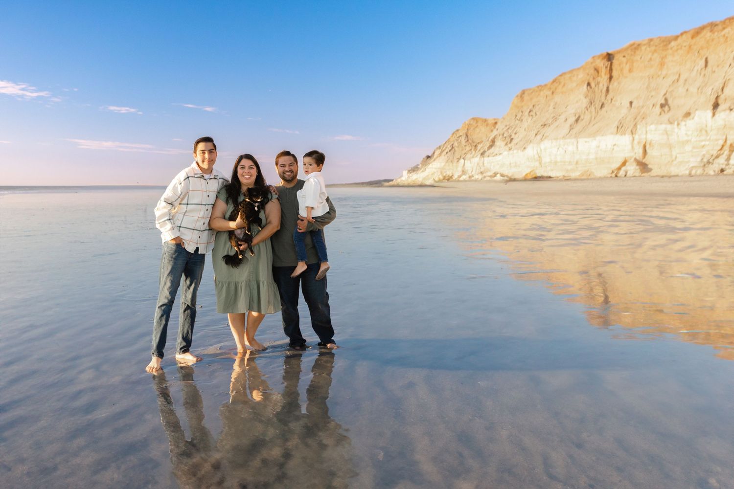 Friends posing together on wet sand with towering beige cliffs and reflective waters behind them.