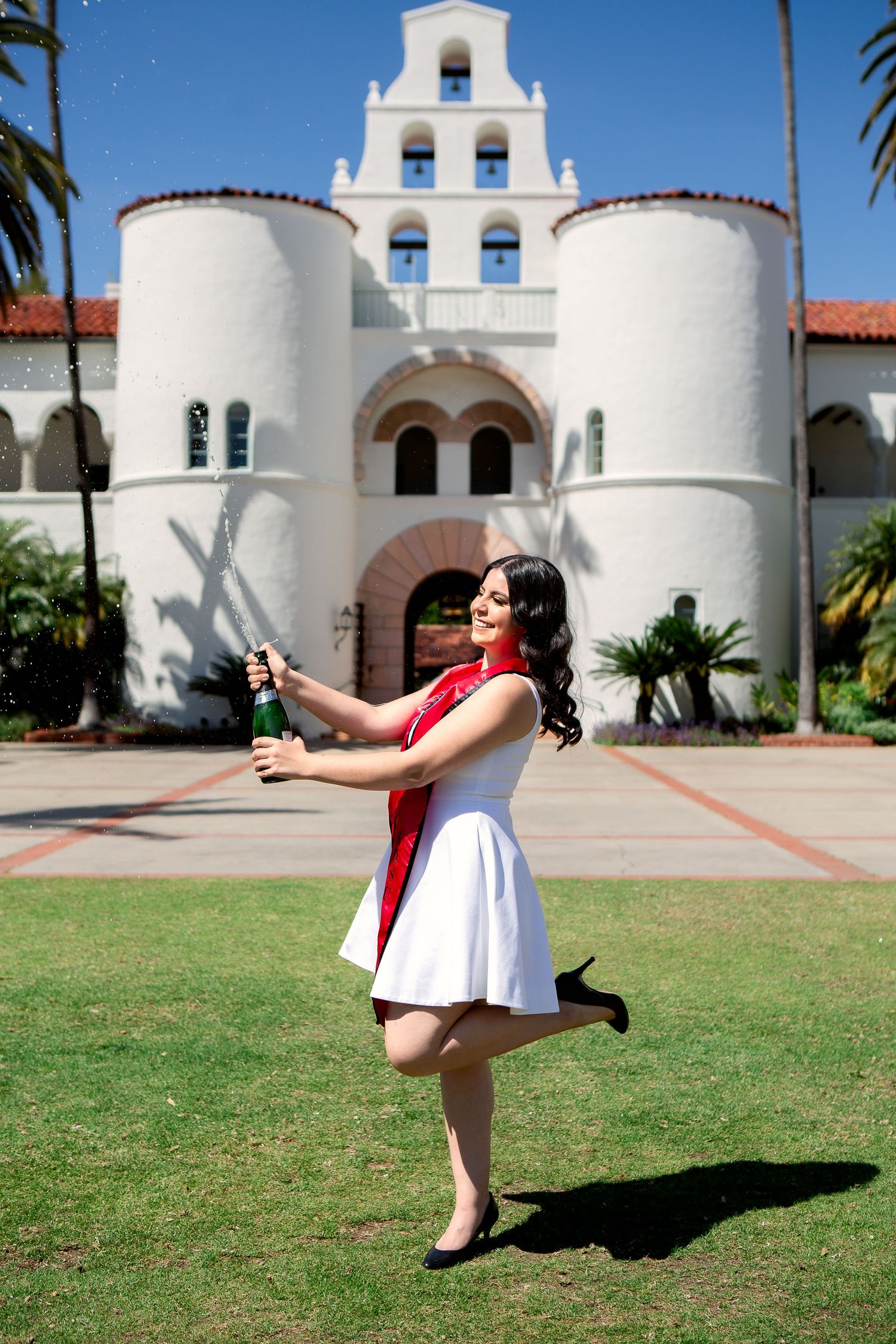 A celebratory graduate poses playfully in a white dress with red sash in front of a Spanish colonial style building.