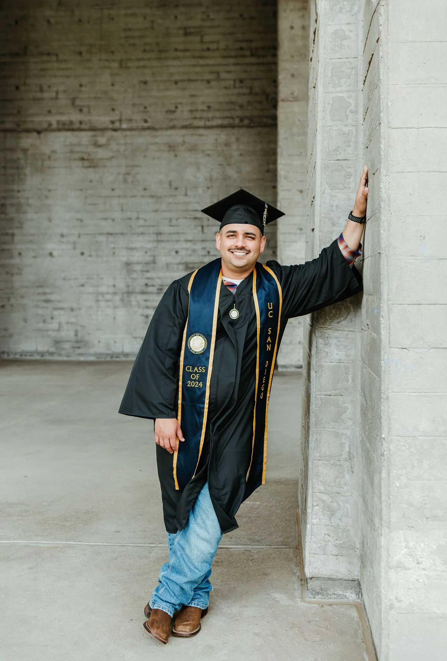 Graduate in black cap and gown with honors cords leans against wall in casual pose.