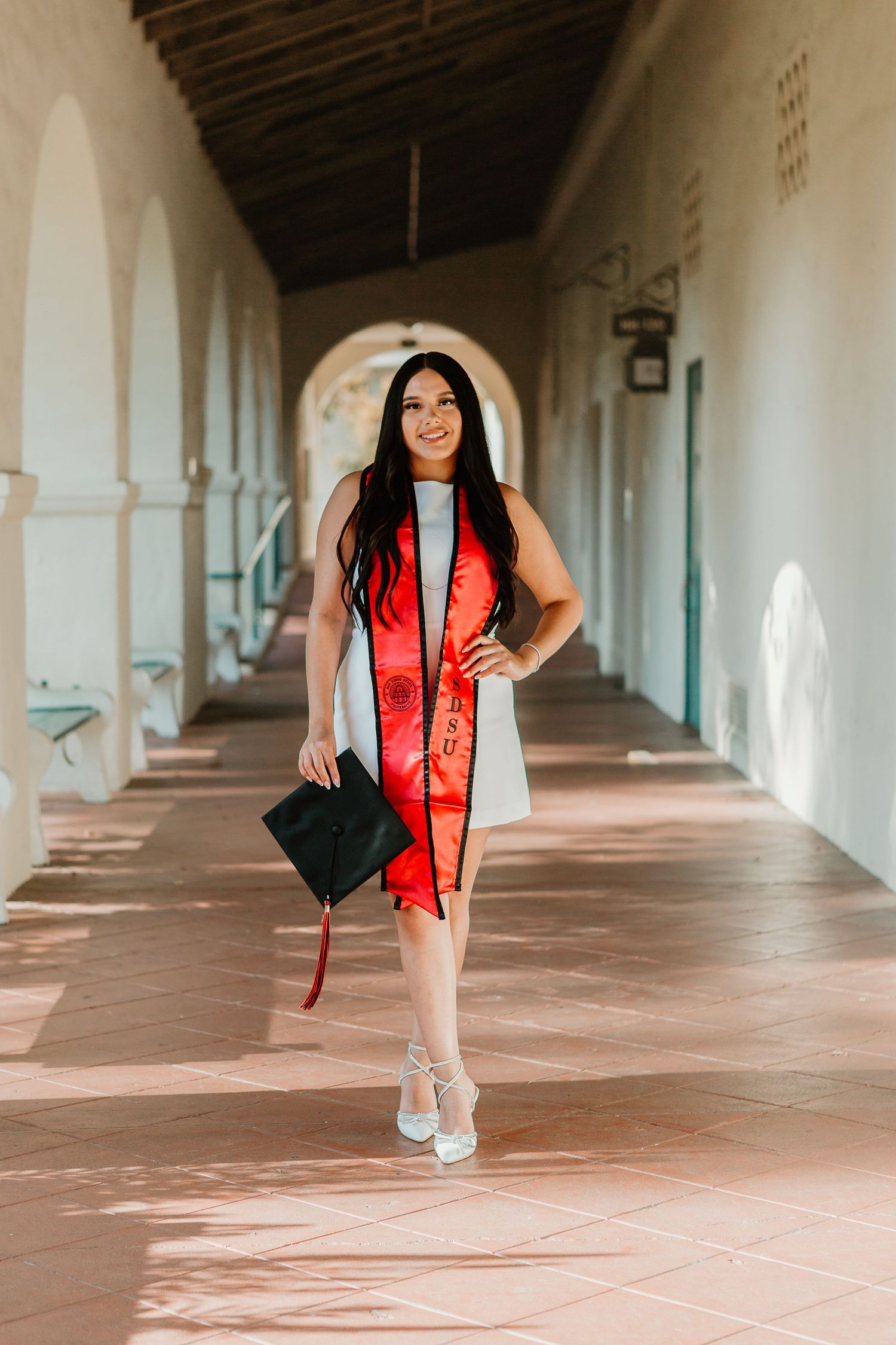 Graduate walks through Spanish colonial archway in white dress with red stole.