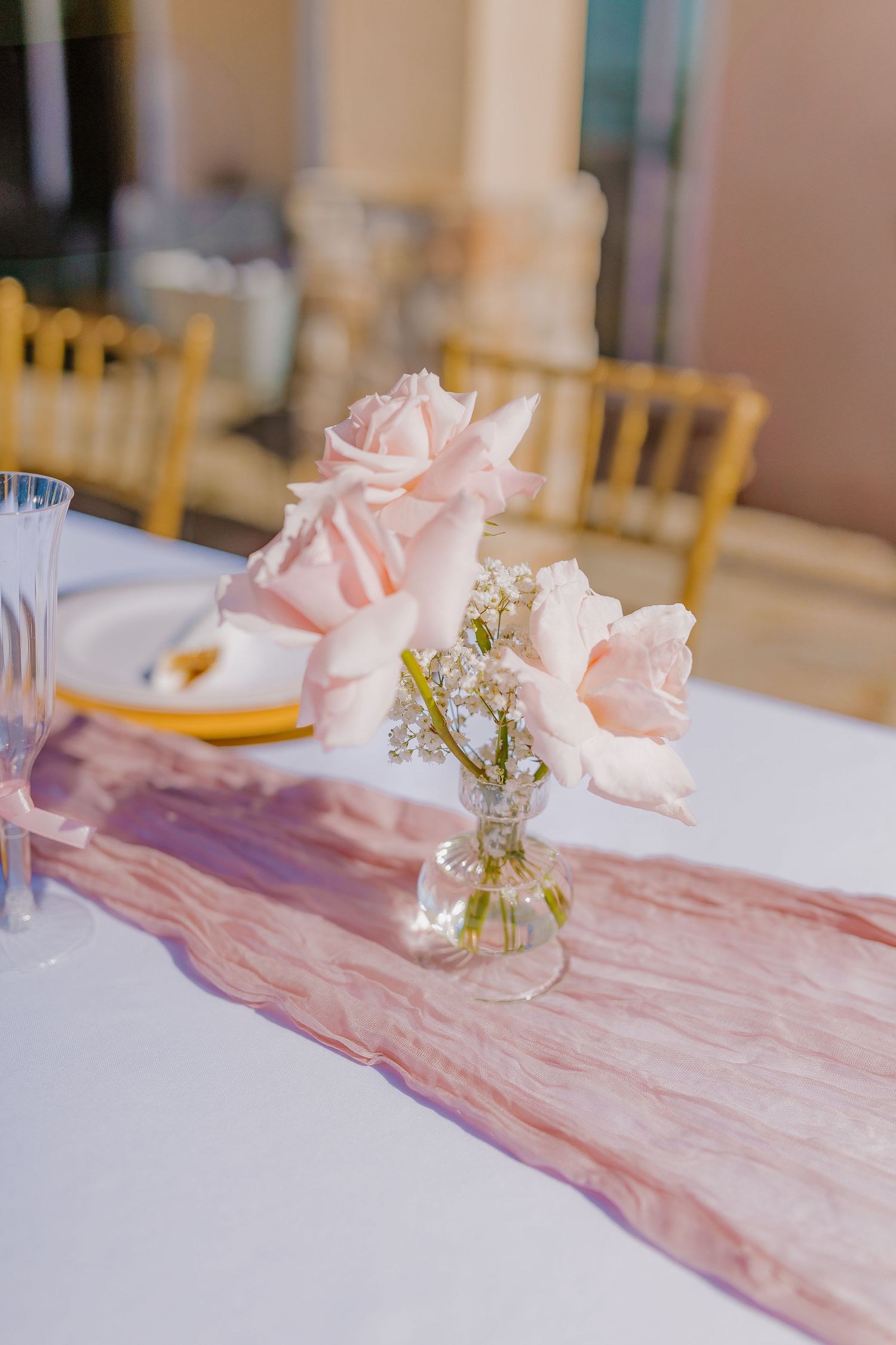 Pink roses in clear vase with delicate table runner on white tablecloth.