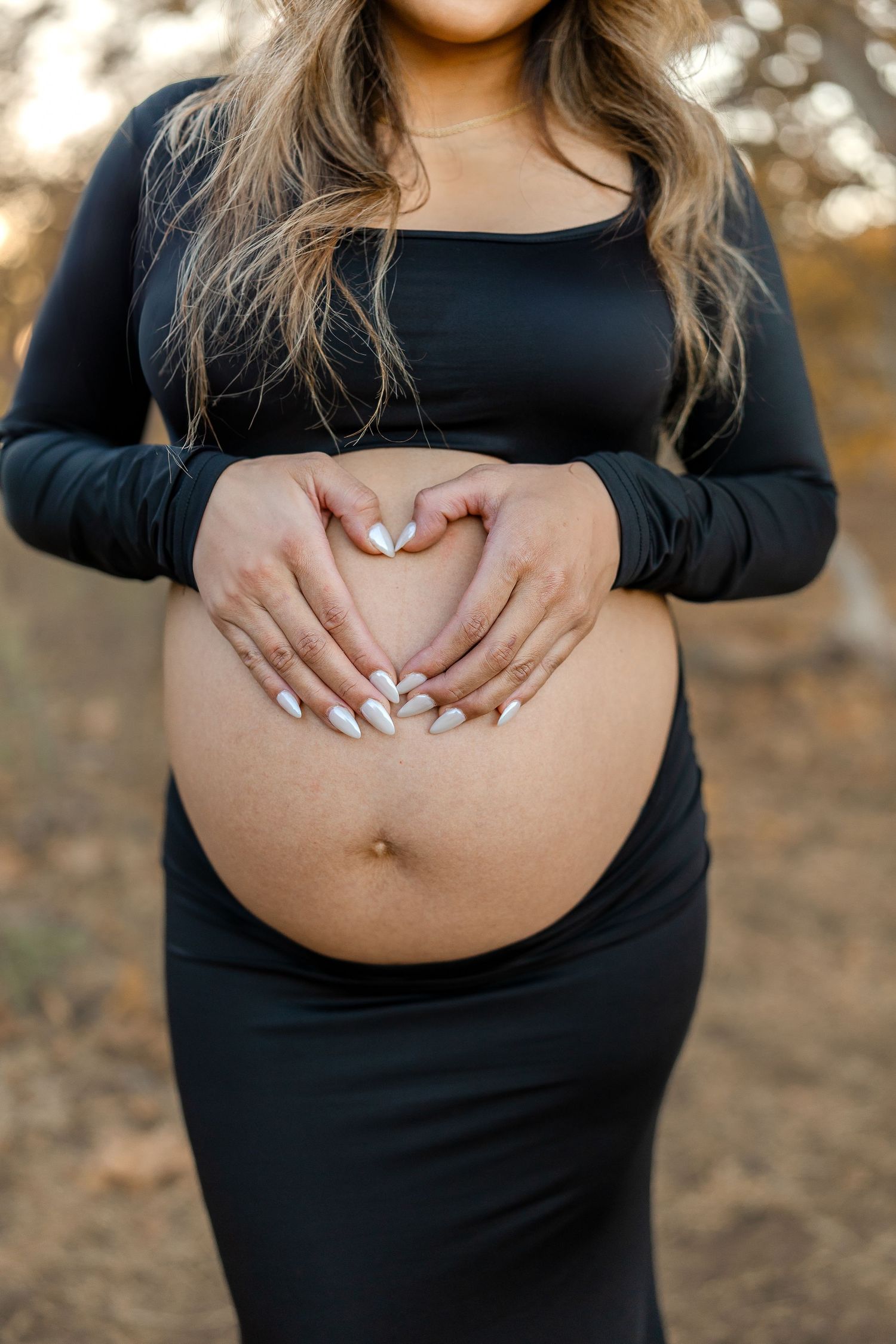 Hands forming heart shape on pregnant belly wearing black dress against natural outdoor background.