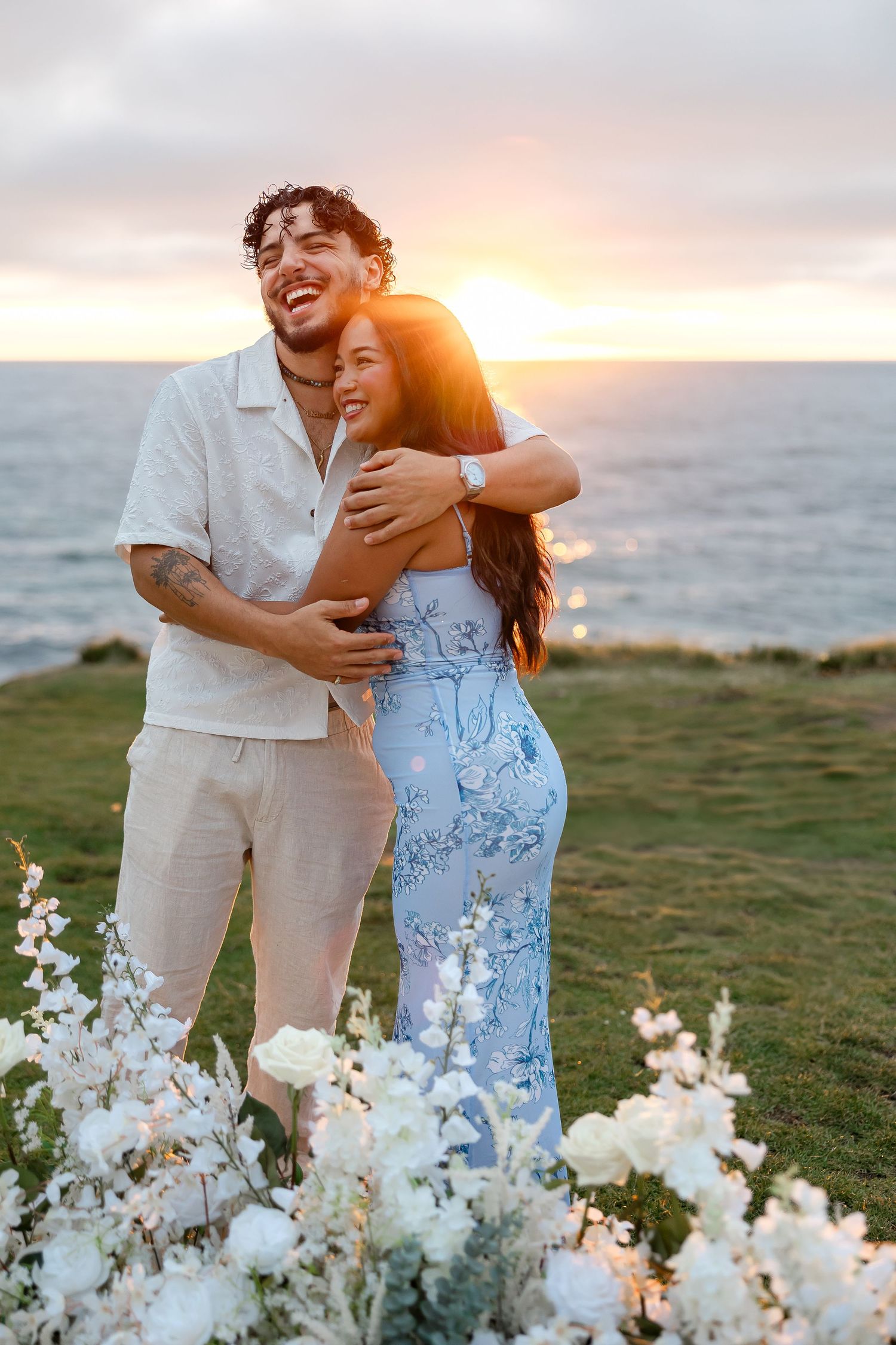 A romantic couple embraces outdoors at sunset with white wildflowers in the foreground on a grassy hillside.