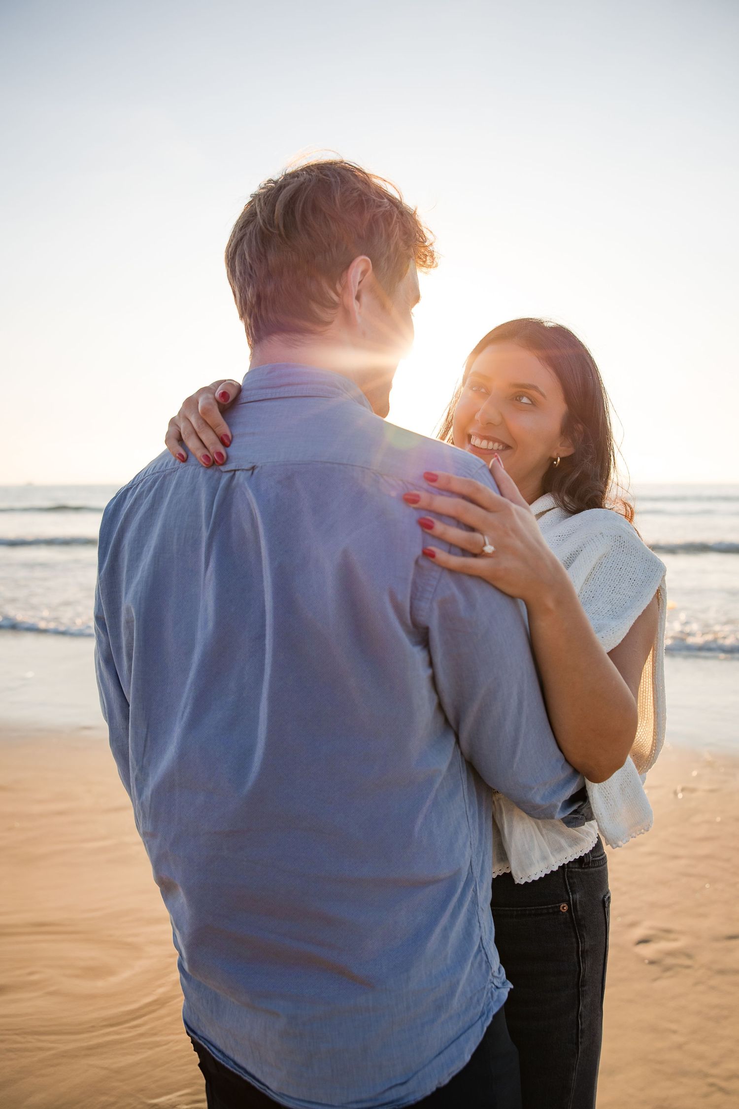 A couple embraces on a sunset beach with golden sunlight streaming between them as waves roll in the background.