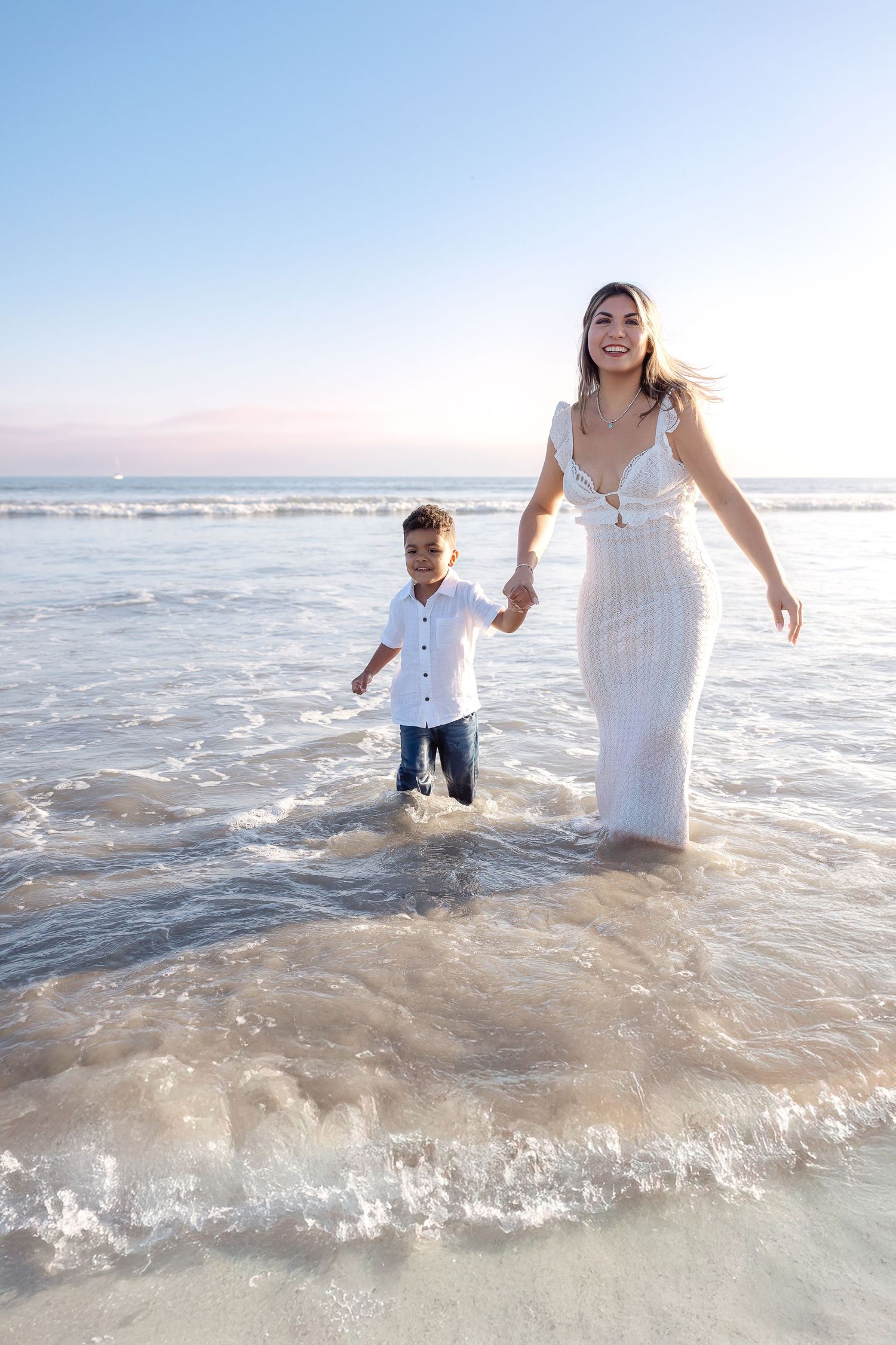 A parent and child wade through ocean waves at sunset, wearing white clothing on a sandy beach.