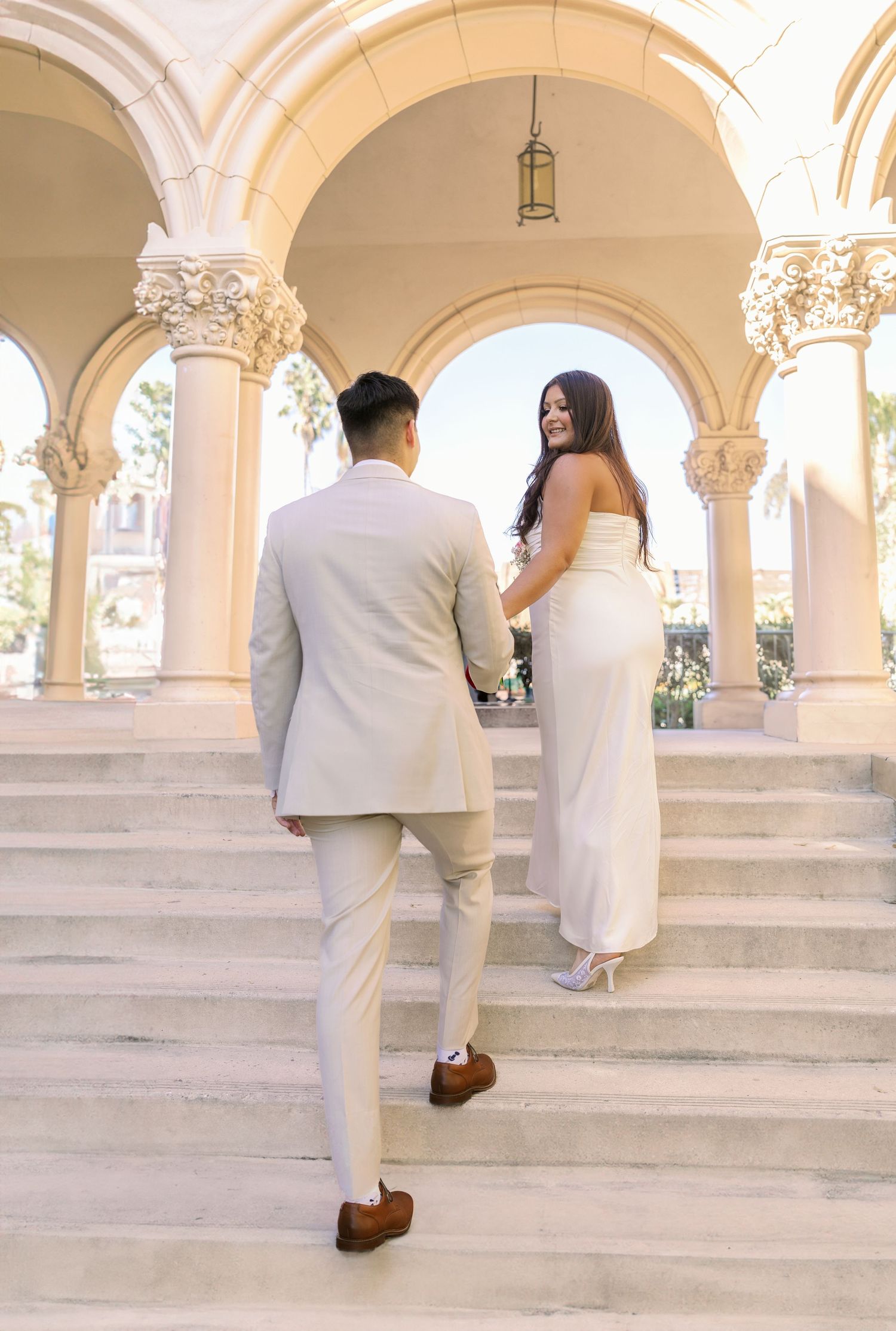 Couple in elegant white attire ascending ornate stone steps with arched columns.