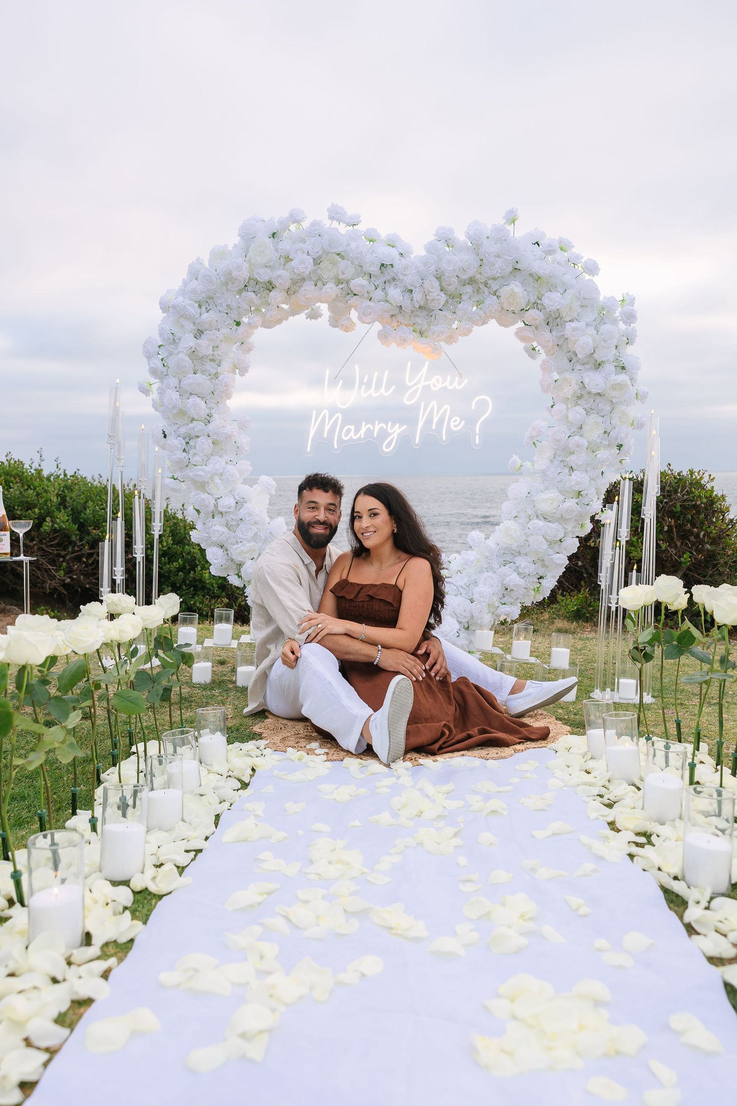 A romantic proposal setup with white floral heart arch, candles and decorated aisle on a scenic clifftop.