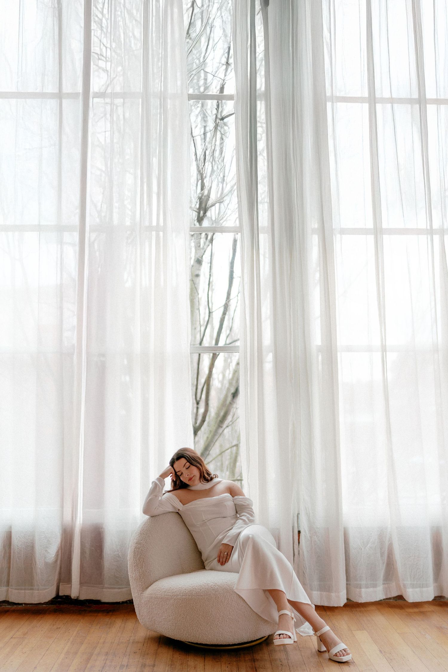White decor interior with sheer curtains and modern upholstered swivel chair on hardwood floors near tall windows.