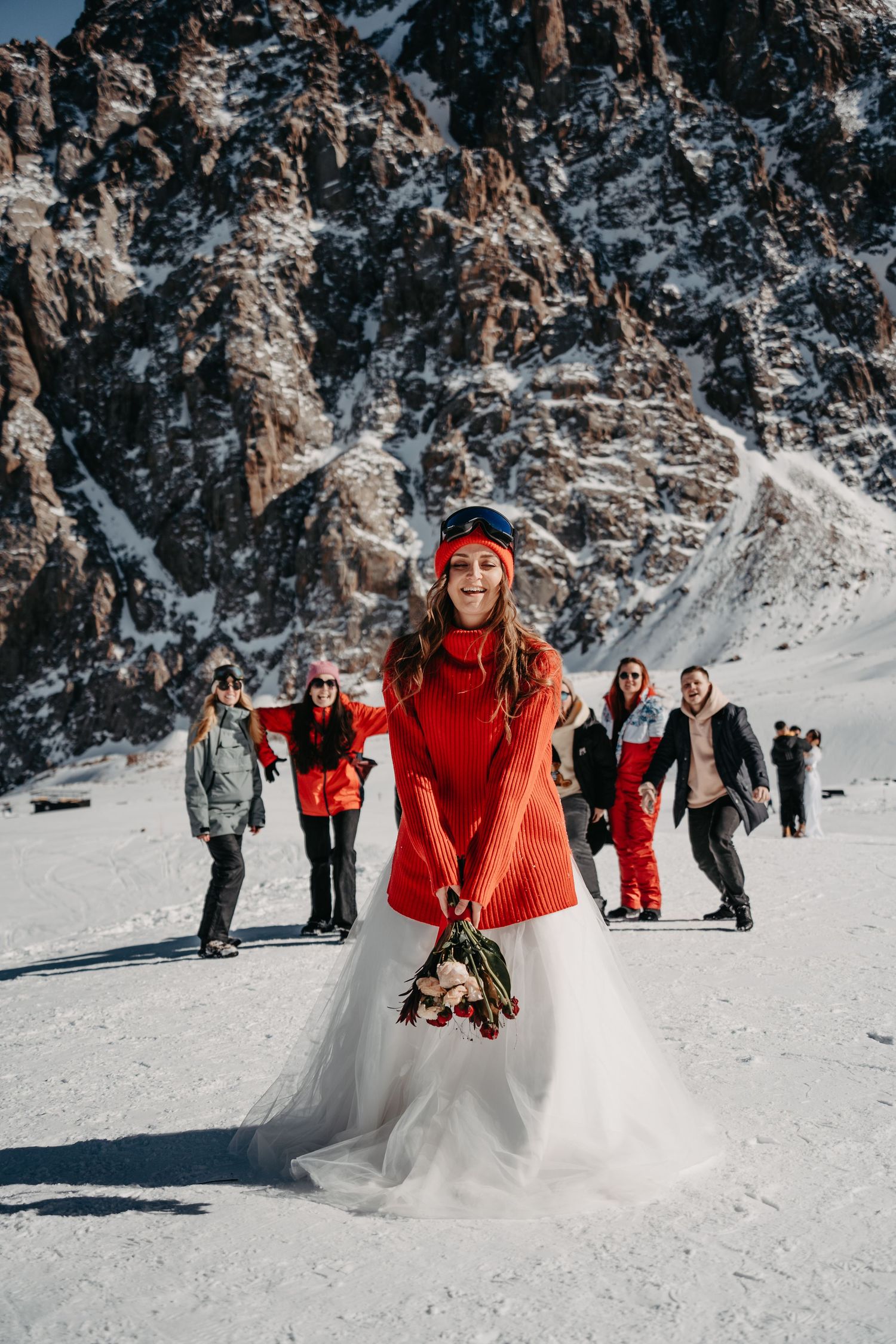 Bride wearing a red sweater tosses her wedding bouquet amidst the snowy mountains