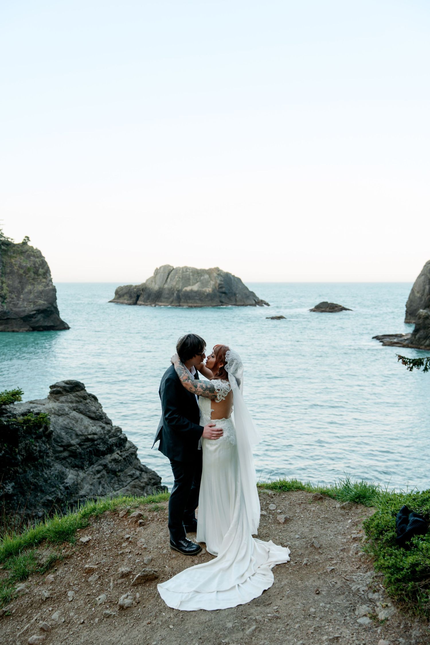 A couple shares a romantic moment on a coastal cliff overlooking rocky outcrops and turquoise waters on their wedding day.