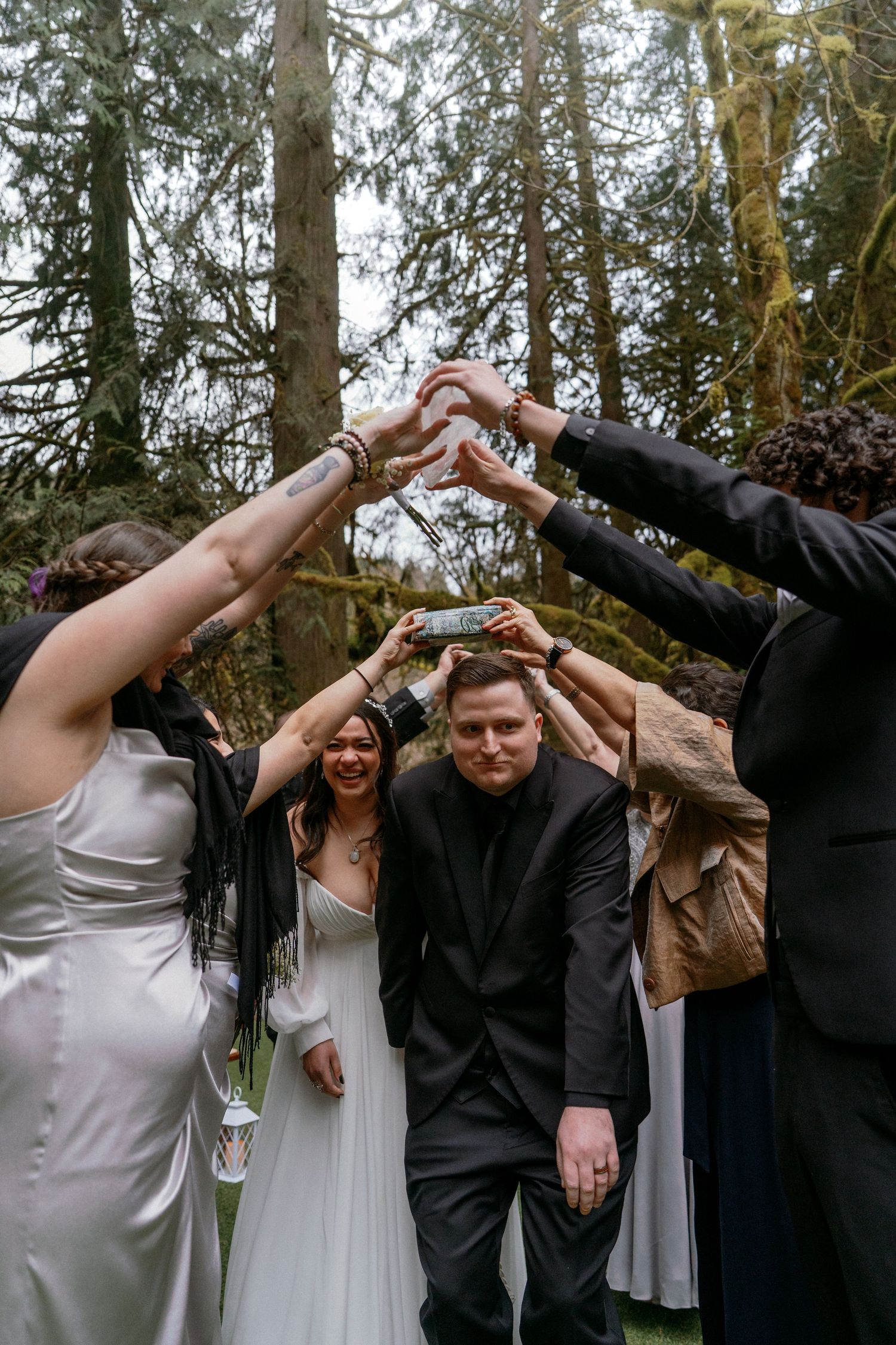 Wedding party forms a tunnel with raised arms in a lush forest setting as guests pass underneath during celebration.