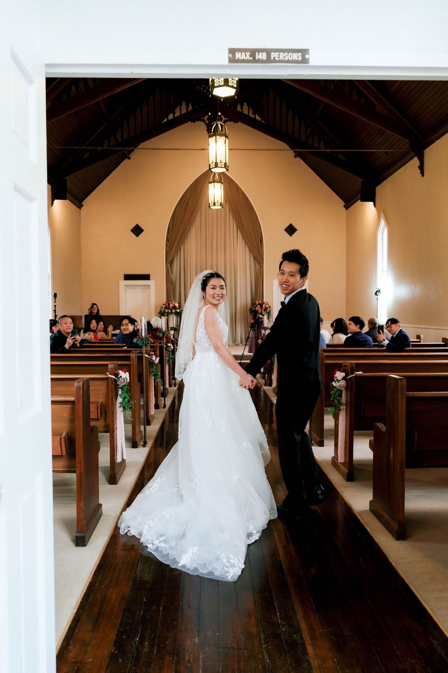 A couple in wedding attire stands together in the aisle of a traditional church with wooden pews and arched doorways.