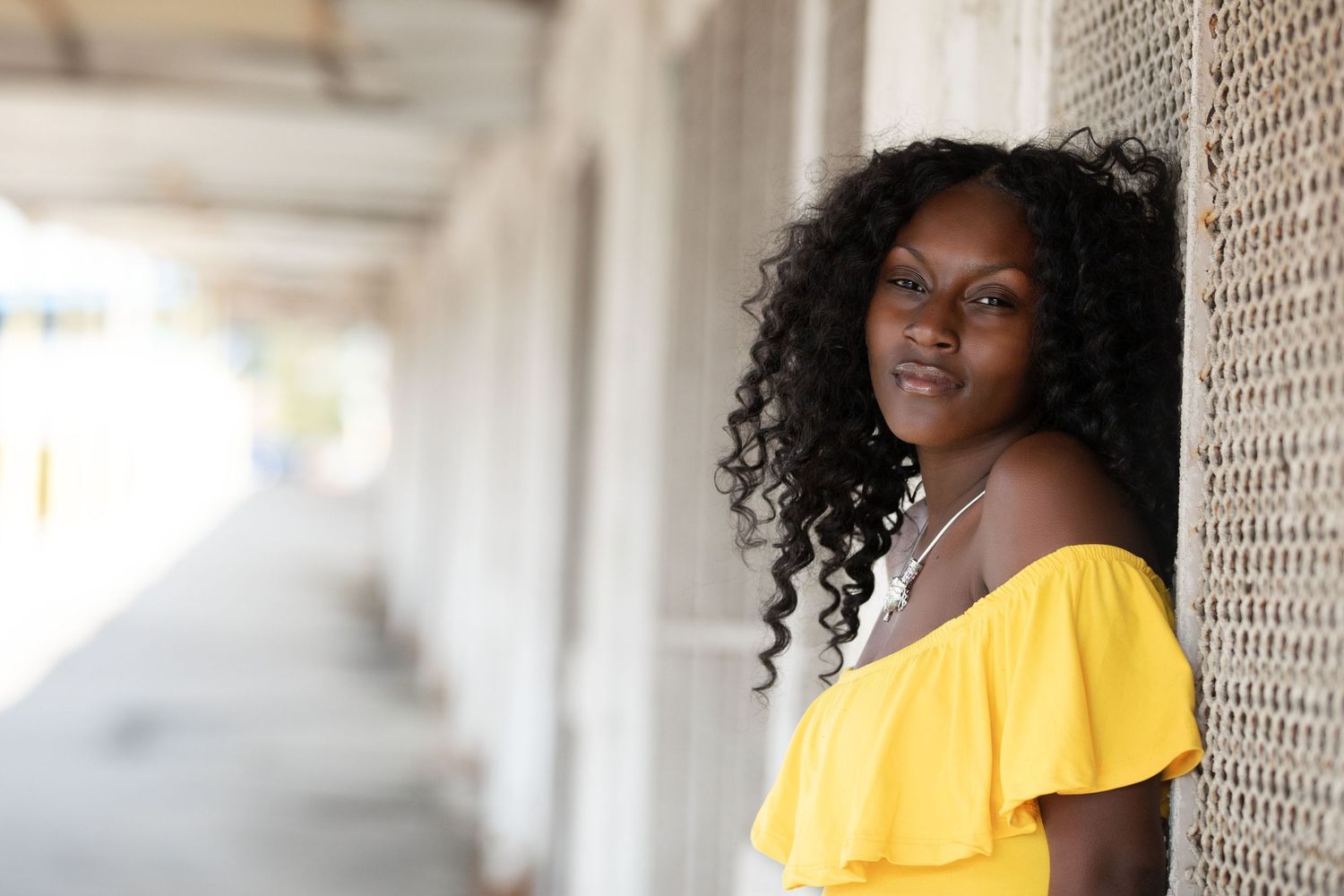 Young Black woman in yellow off-shoulder top leaning against wall, smiling confidently in outdoor corridor.
