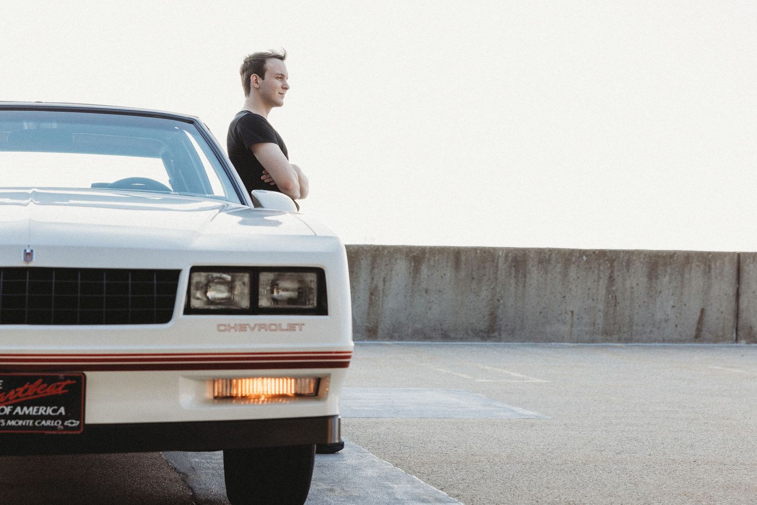 Man leaning against a white Chevrolet Monte Carlo in a parking lot, casual pose with arms crossed.