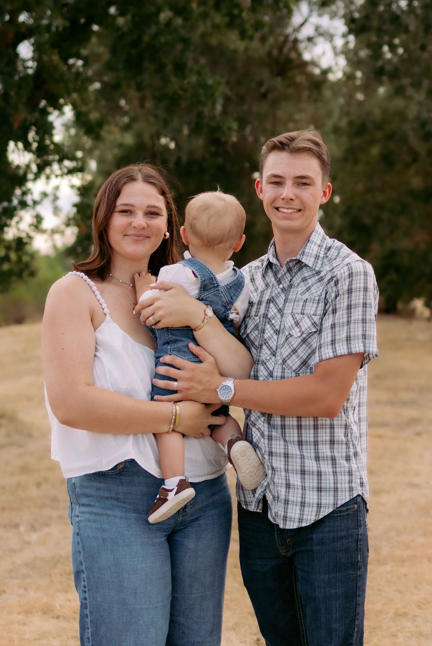 Young couple smiling and holding baby in denim overalls in sunny park setting.