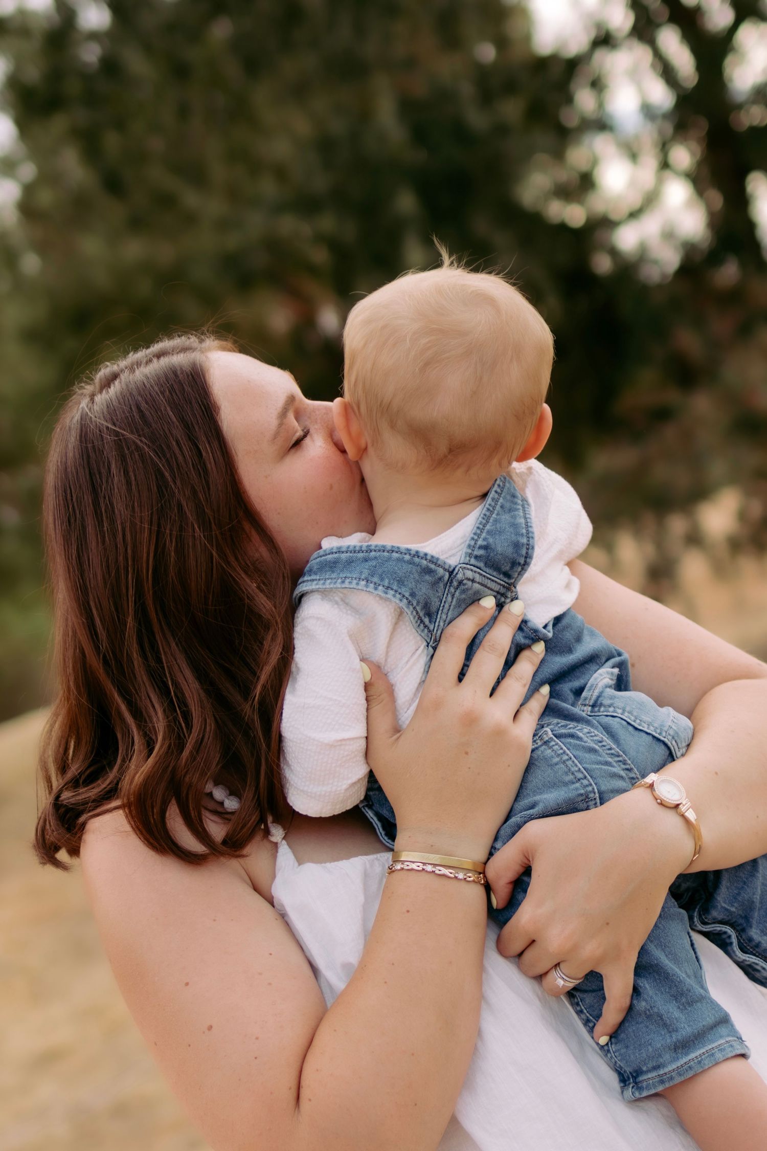 Loving mother kissing baby in denim overalls while holding them in outdoor park.