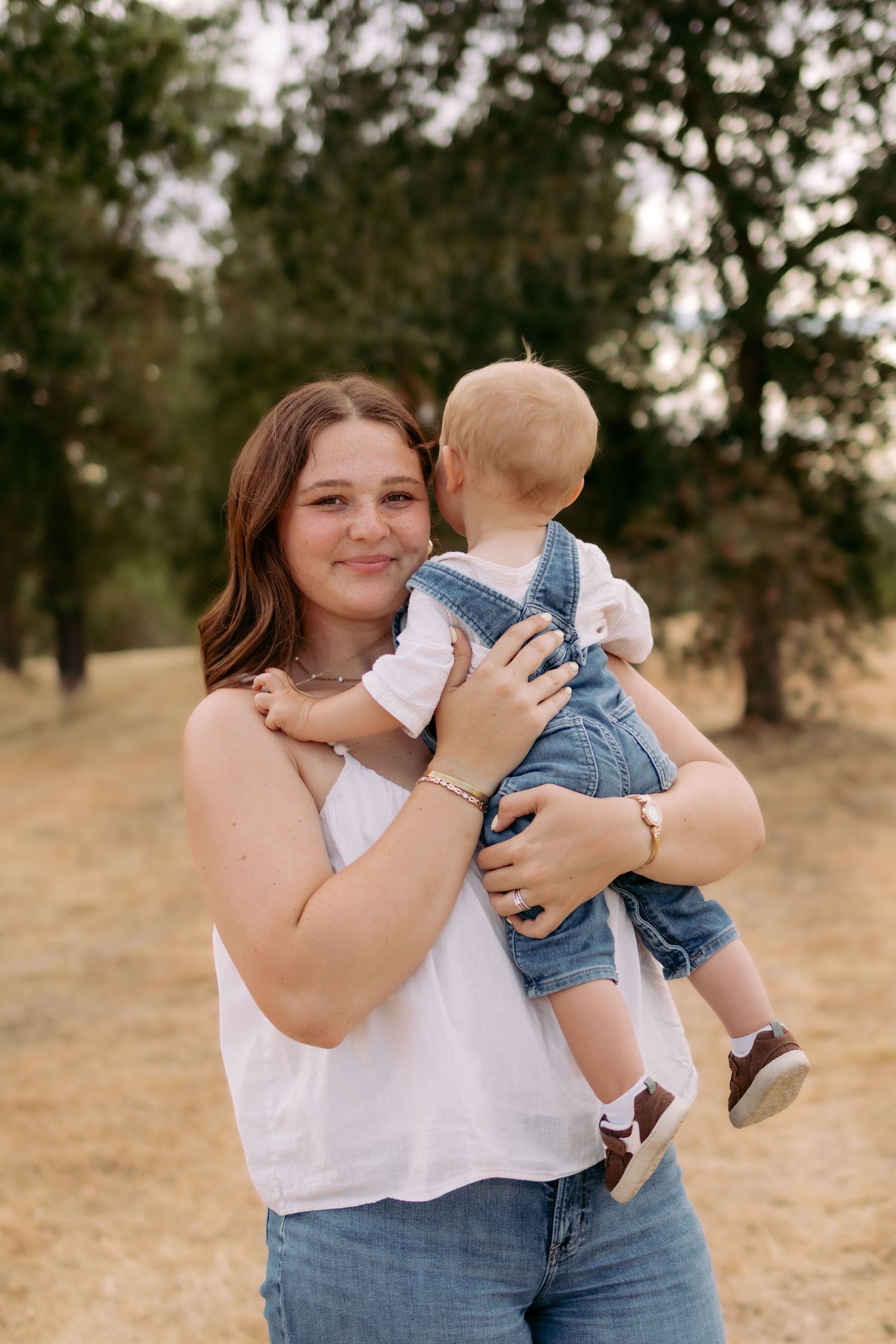 Smiling brunette mother holding baby in denim overalls in golden hour park setting.