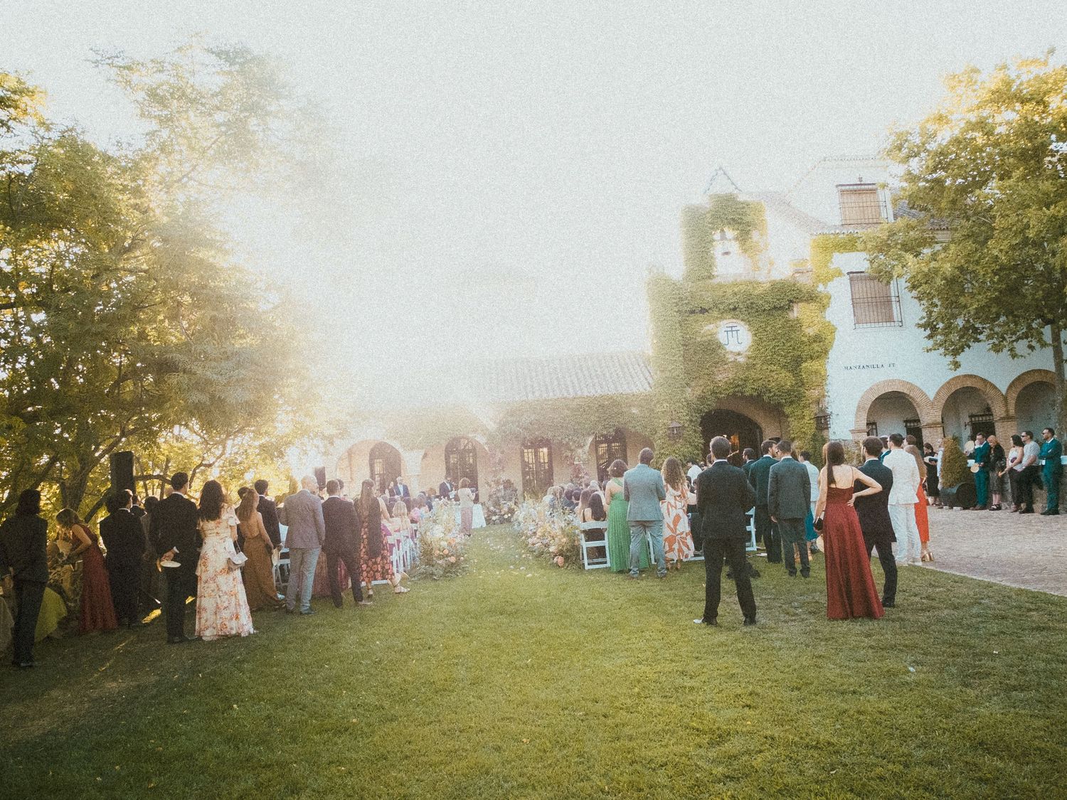 Large group of guests gathered outdoors at a rustic wedding venue during golden hour.