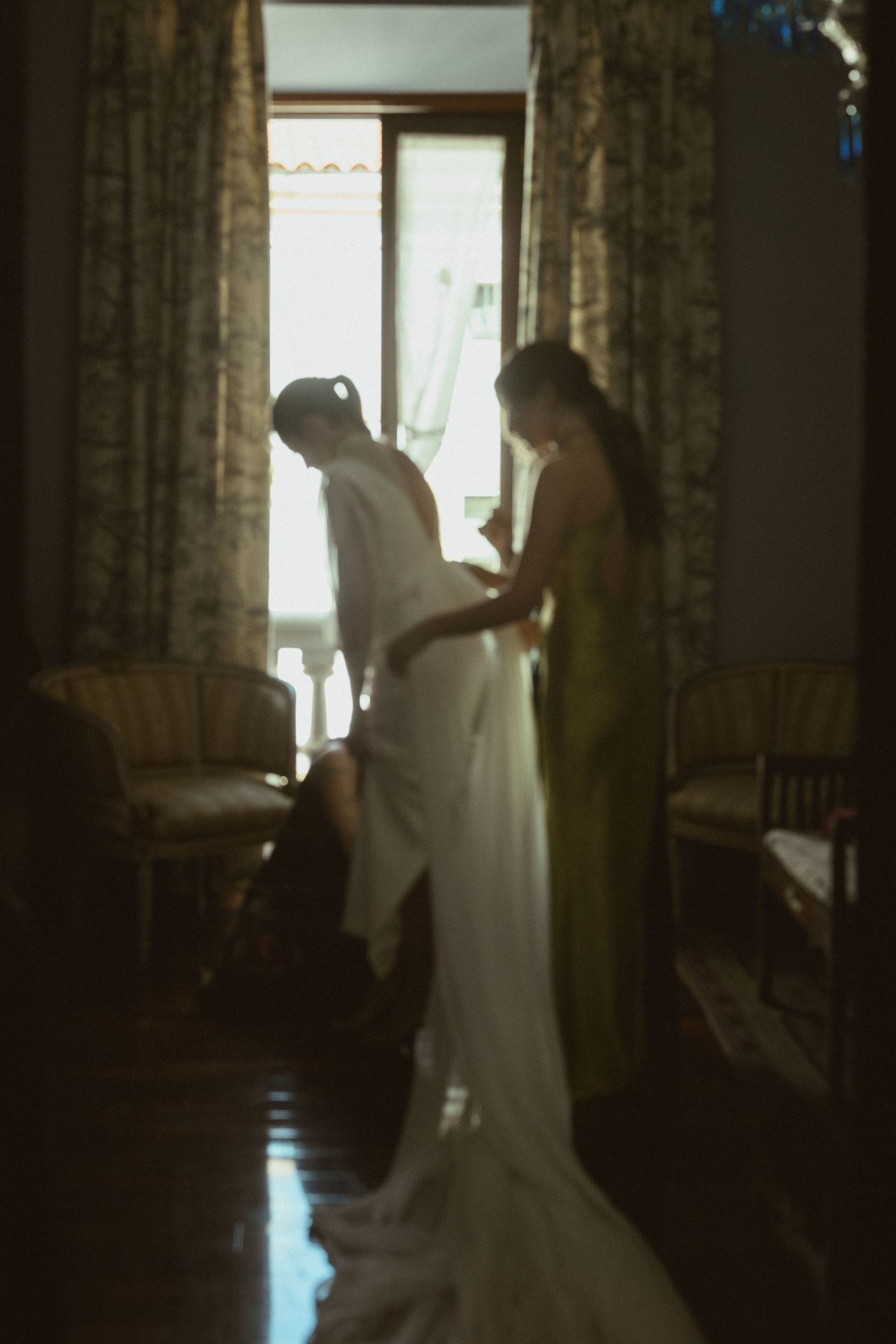 Silhouetted figures prepare in a sunlit room with flowing white fabric and curtains.