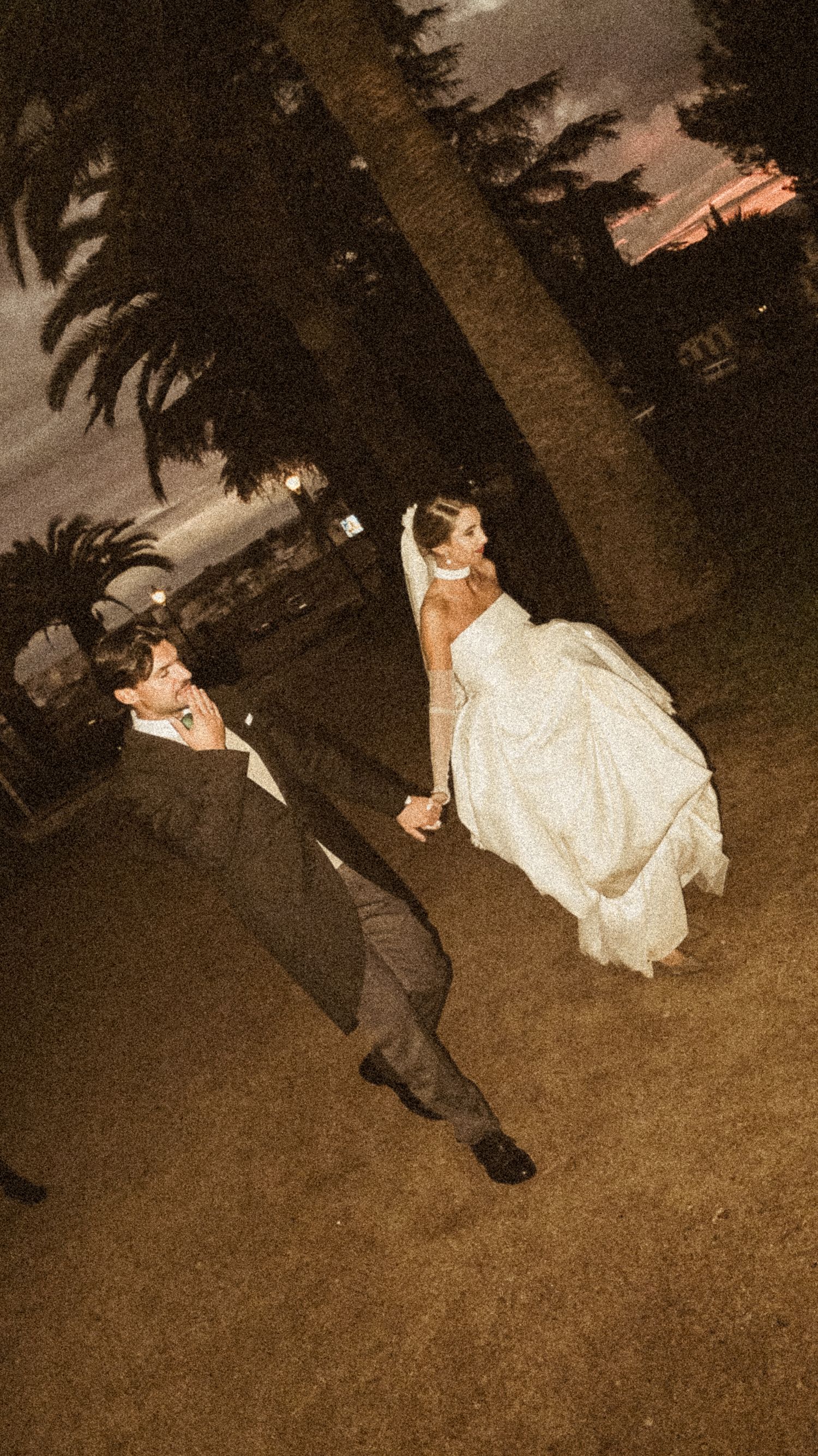 A couple in wedding attire walking under palm trees at night.