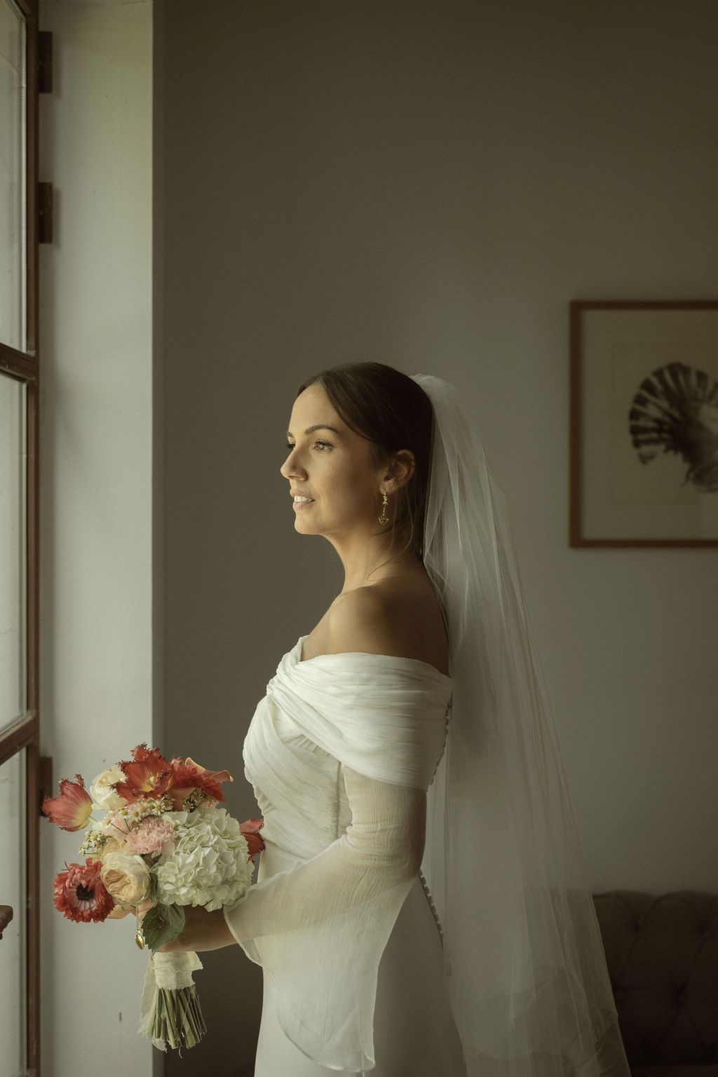 Bride in white dress and veil poses elegantly by window with pink and white bouquet.