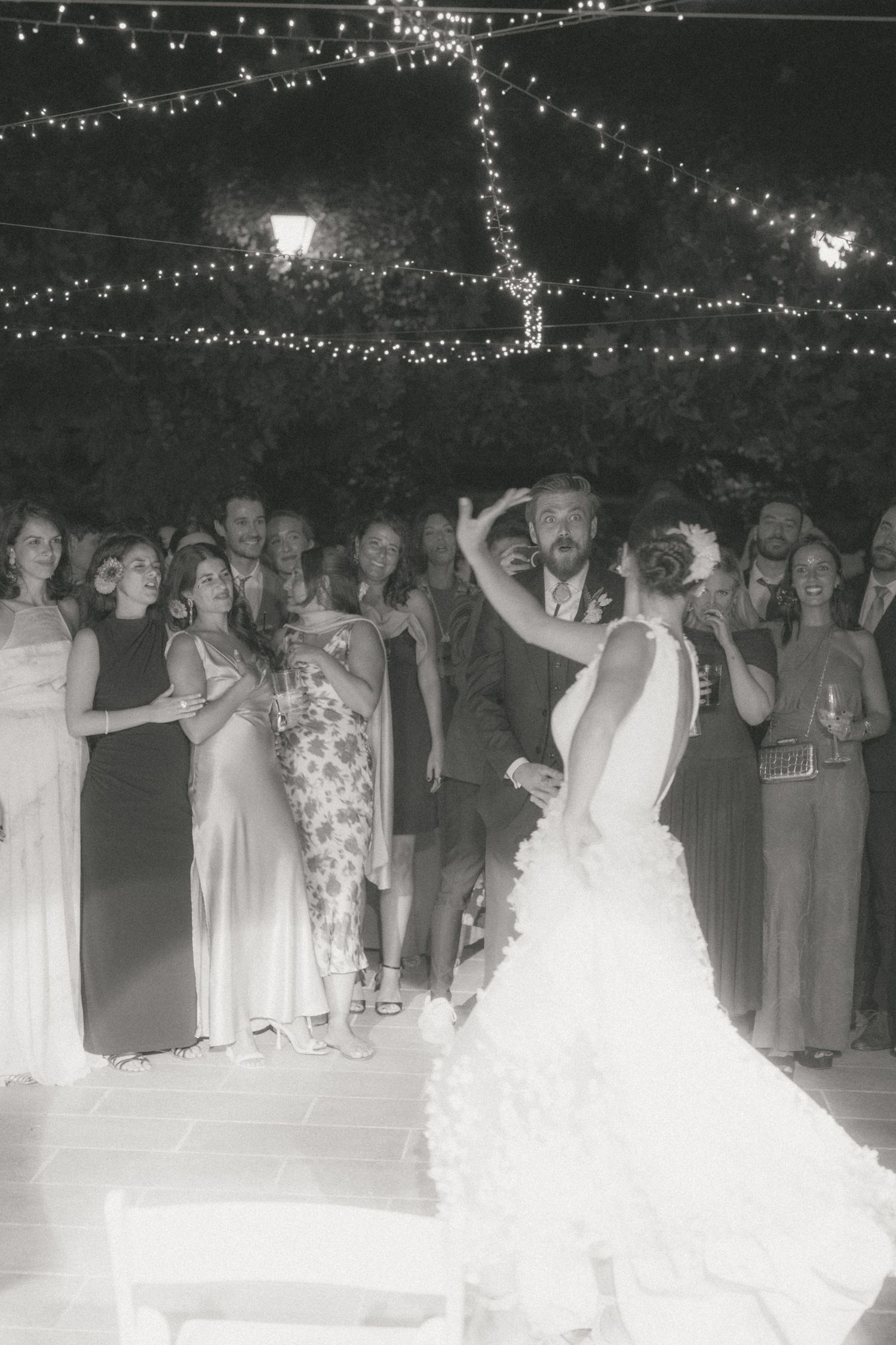 Wedding party and guests dancing under string lights at outdoor reception.