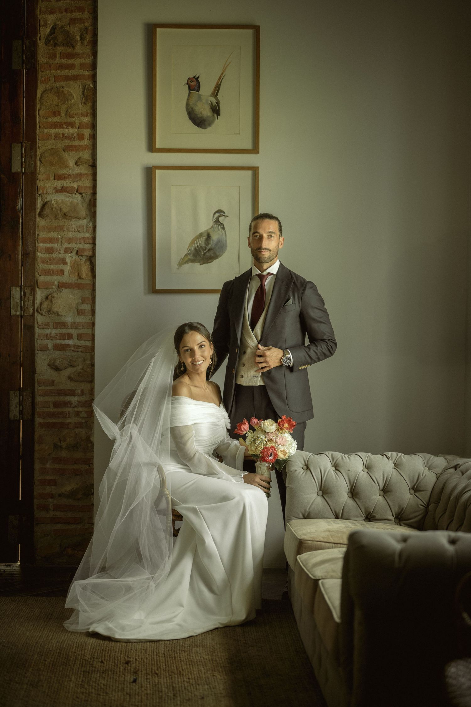 Couple in wedding attire poses on vintage tufted couch in rustic brick room.