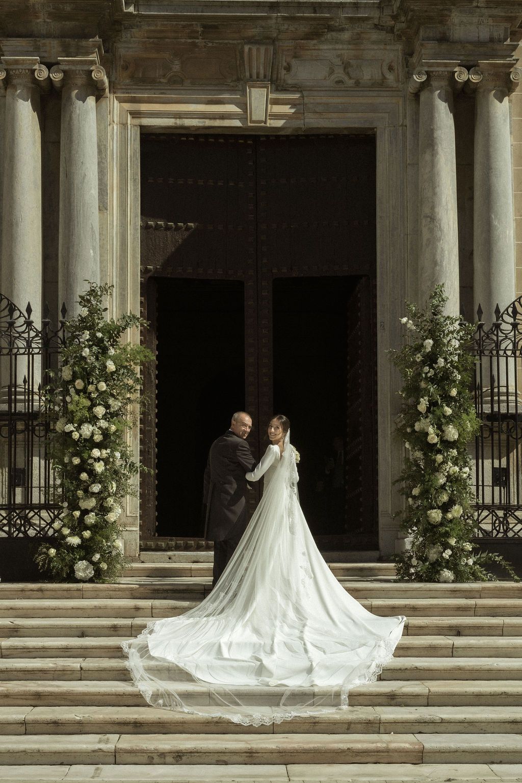 A dramatic wedding photo shoot with white floral arrangements along classical stone columns.