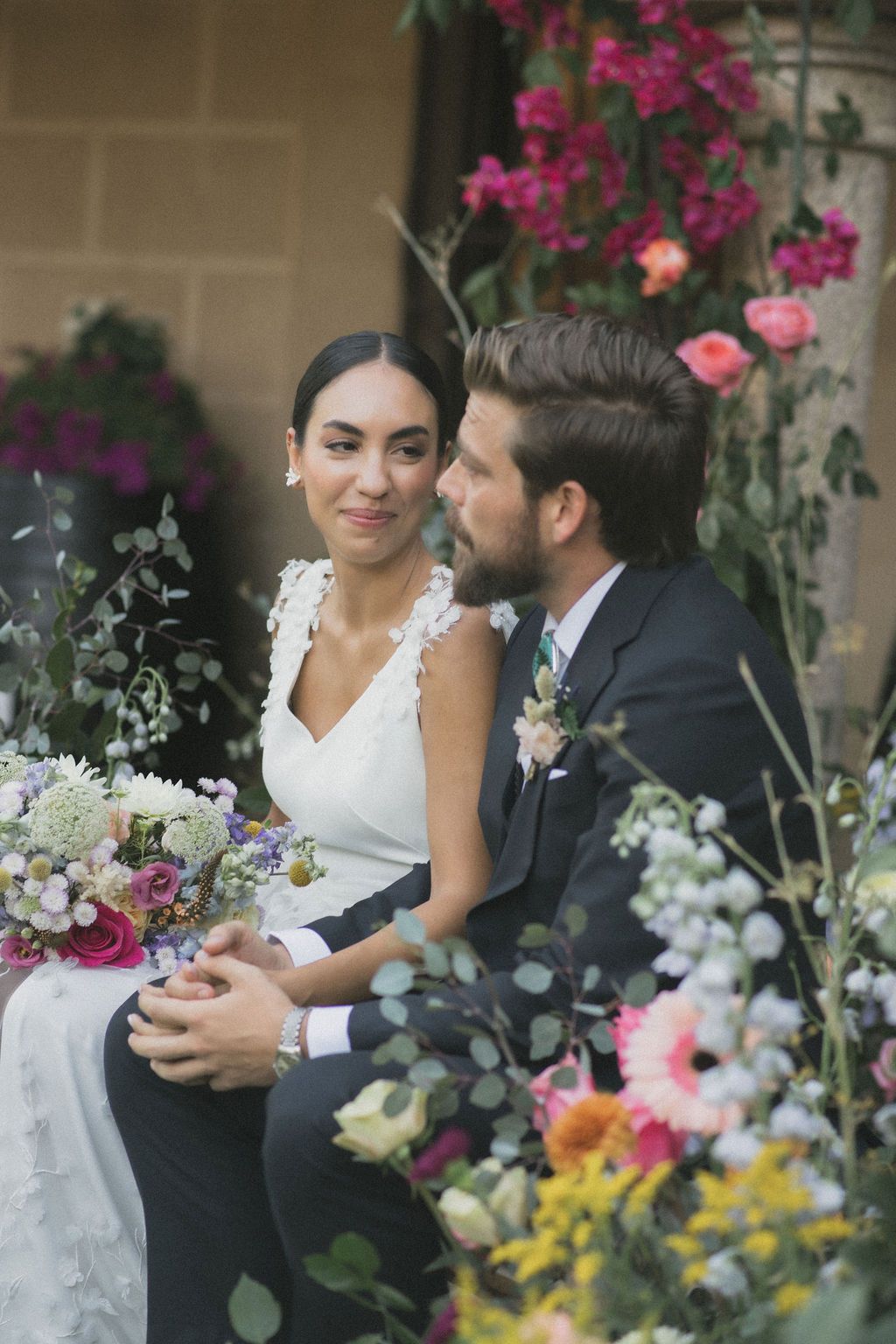 Wedding couple surrounded by colorful floral arrangements in an outdoor garden setting.