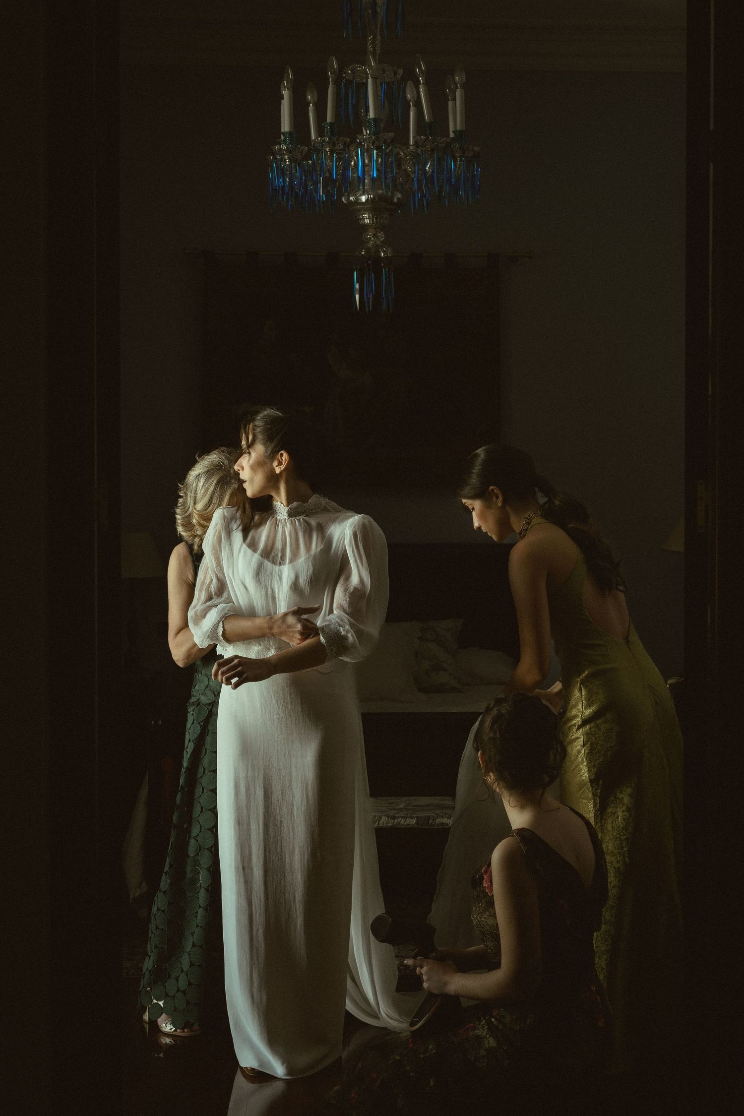 Wedding preparation scene in a dimly lit room with a chandelier and elegant white dress.