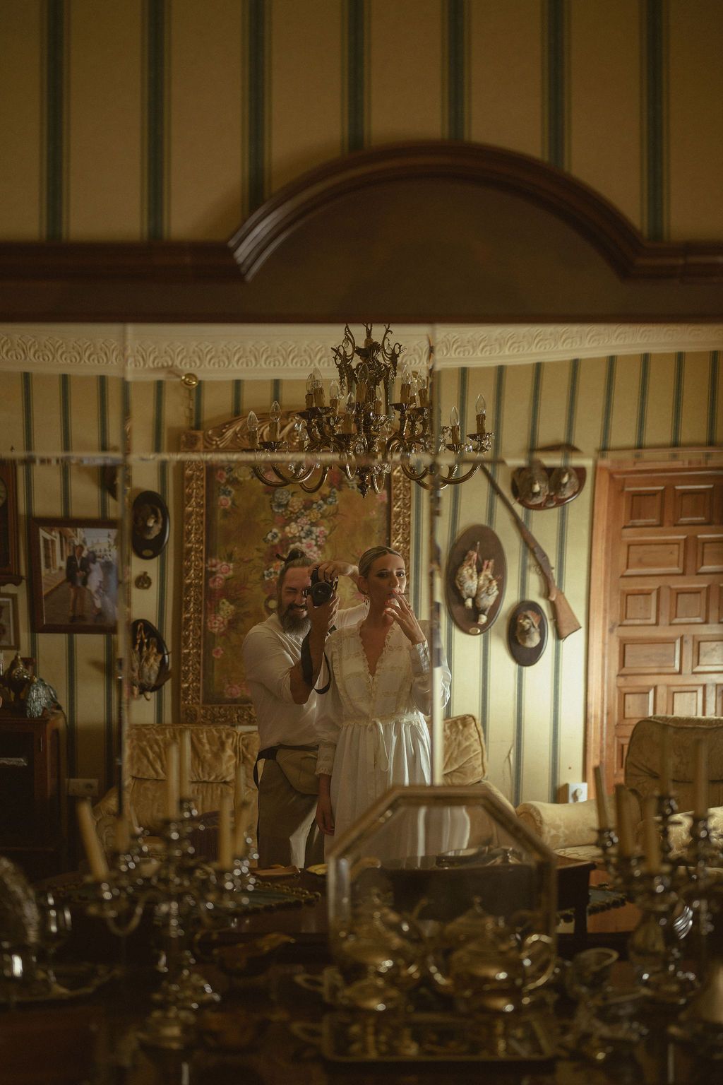 Reflection in an ornate mirror shows a couple in formal attire standing beneath an elegant chandelier.